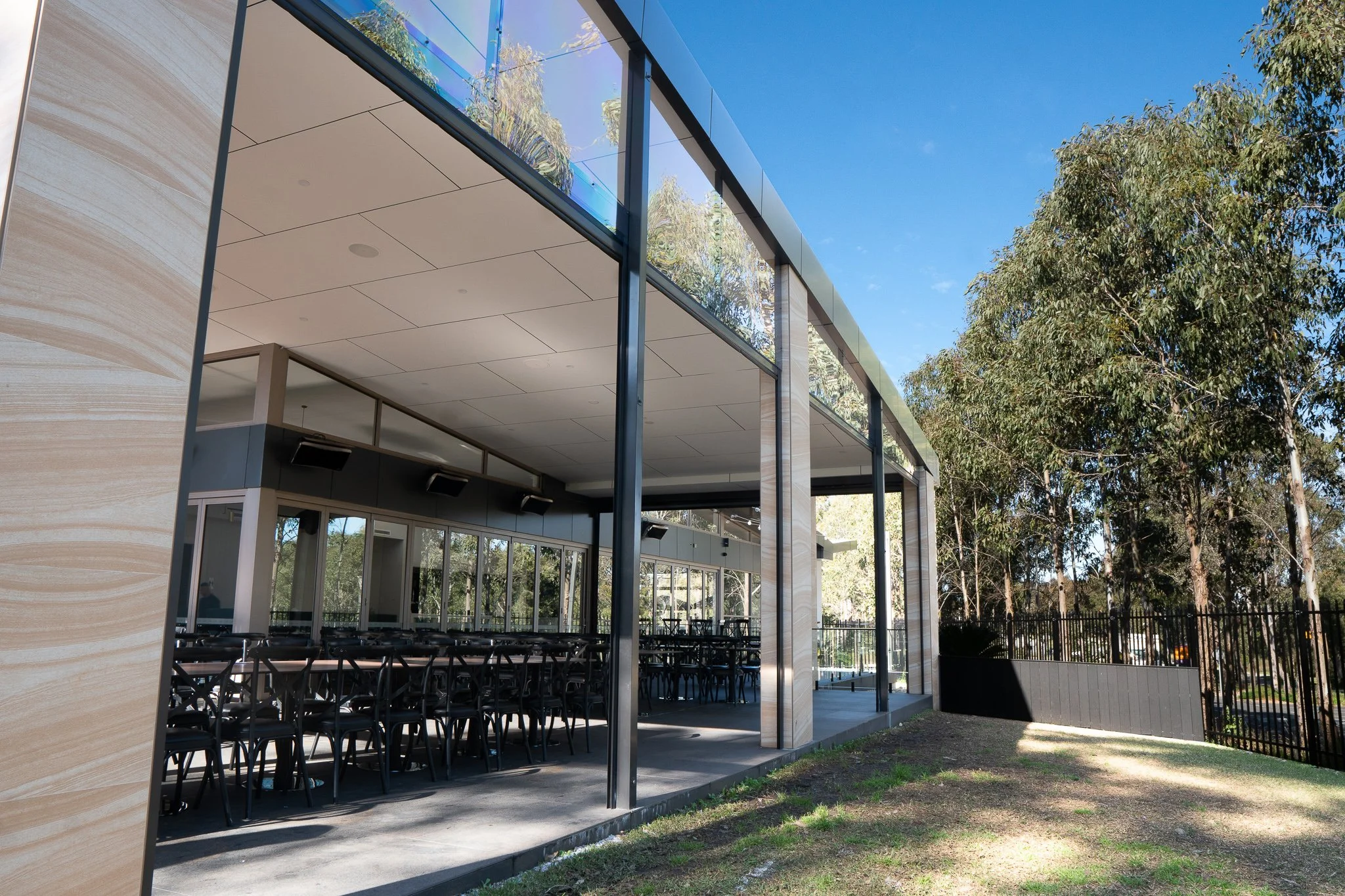 Modern building with large glass windows and an outside view of trees, featuring an open-air patio with tables and chairs.