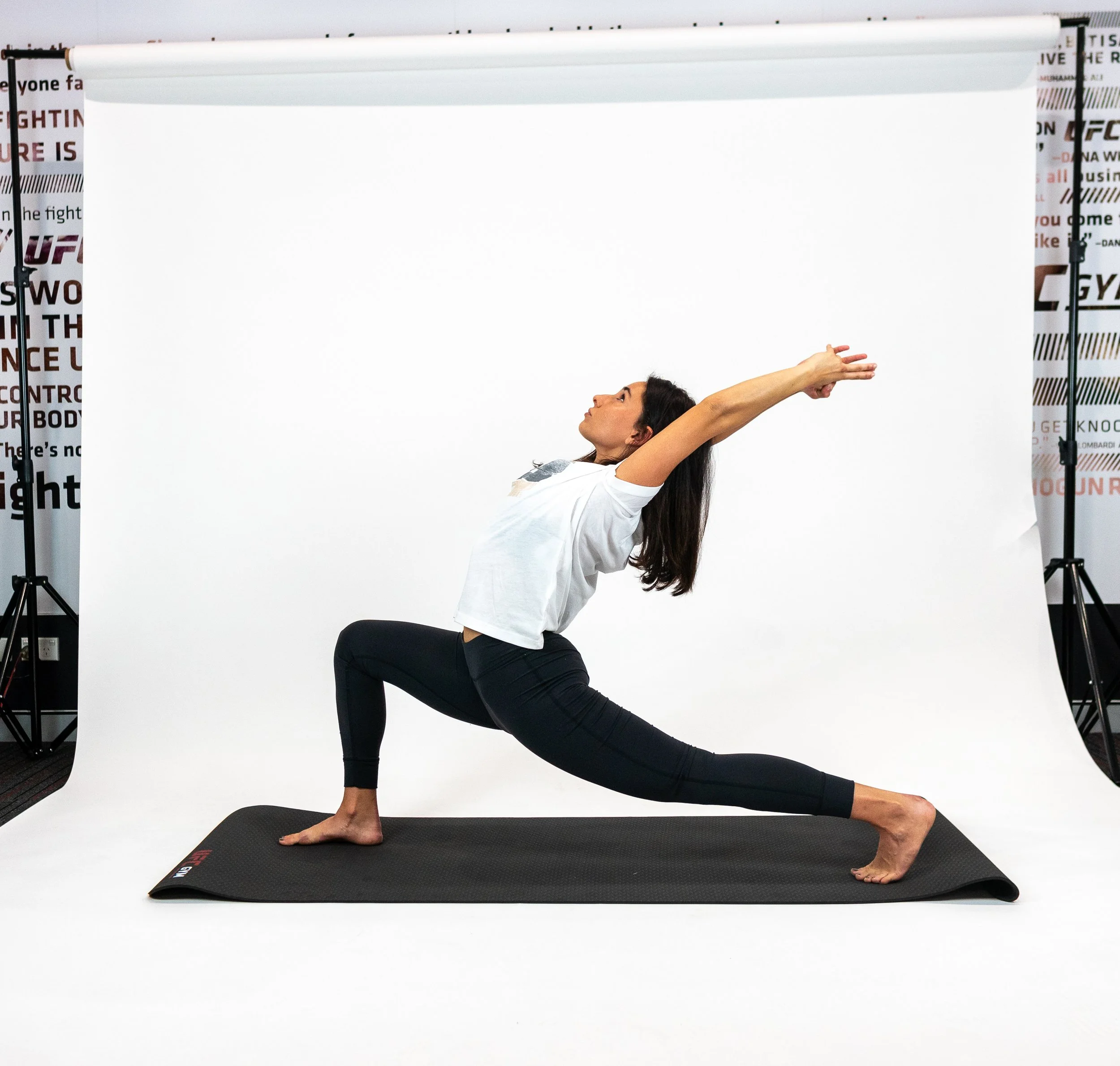 Woman practicing yoga in a lunge pose with arms extended forward in a studio setting with a white backdrop.
