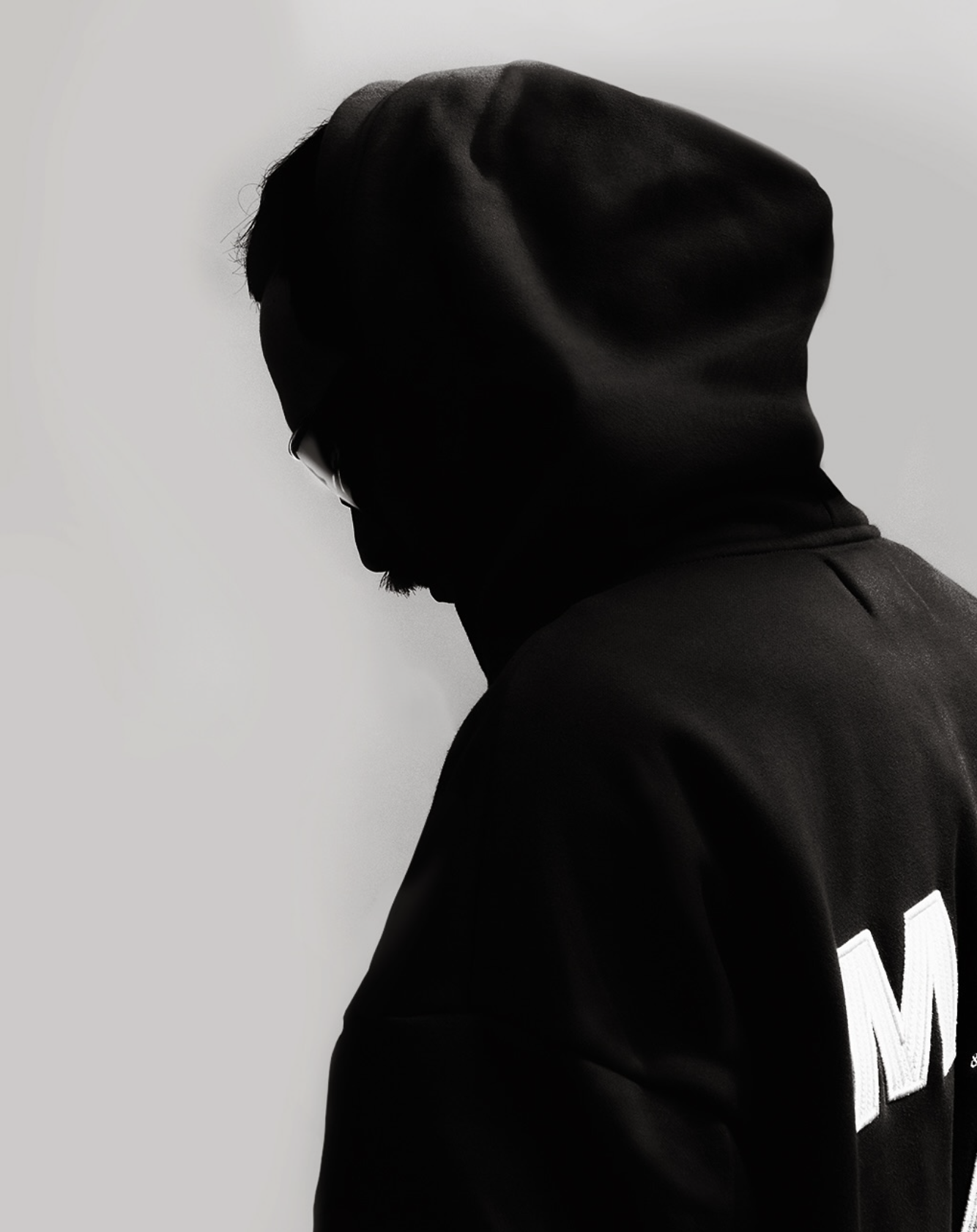 Side profile of a person in black hoodie and jacket with white lettering, facing a plain white wall, with minimal lighting and shadowing, creating a moody silhouette.