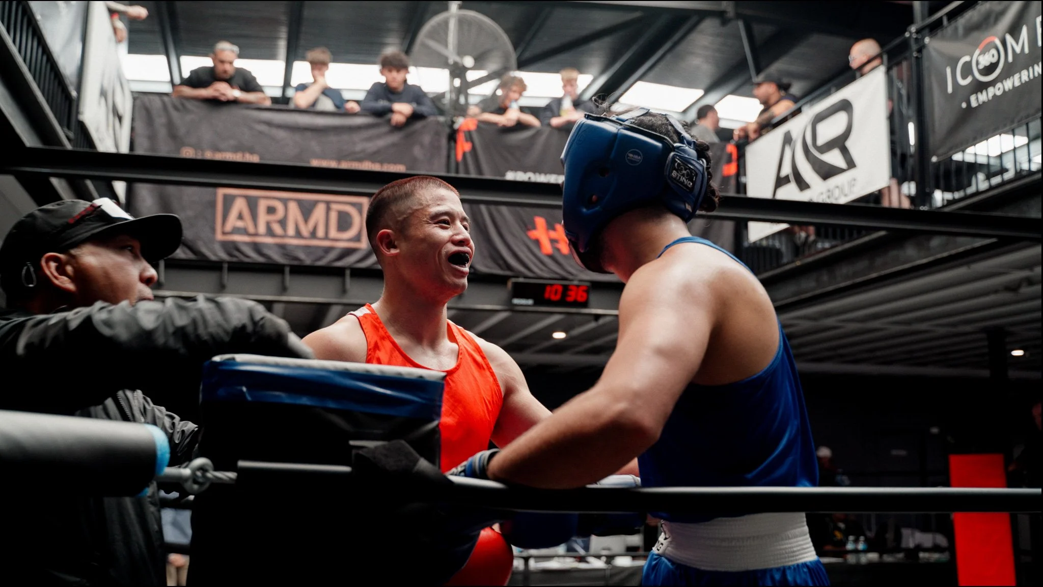 Two male boxers in the boxing ring, engaged in a match. One is wearing a red tank top and is talking or yelling, the other is in a blue tank top and headgear. A referee is partially visible, and spectators are watching from a balcony above.