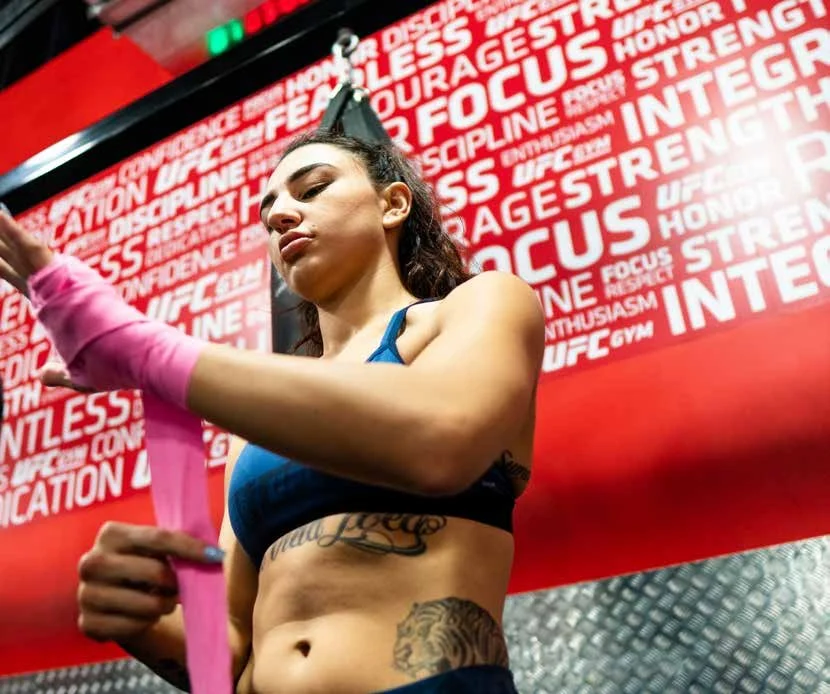 A young woman with tattoos, wearing a sports bra, is preparing a pink resistance band in a gym with motivational words on the red wall behind her.