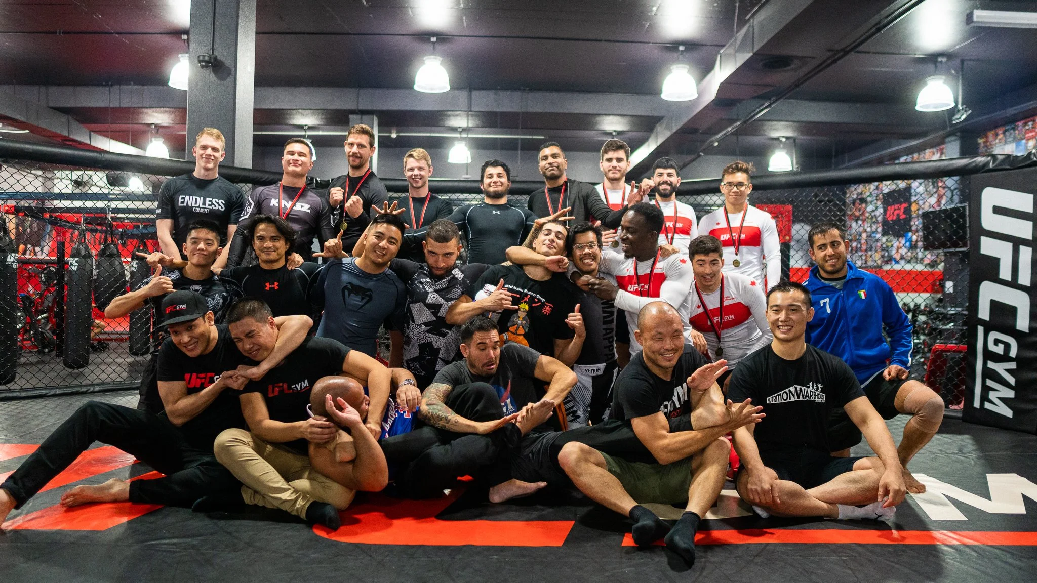 Group of people posing inside a UFC gym octagon with various athletic and casual attire, some wearing medals, and a cage surrounding them.