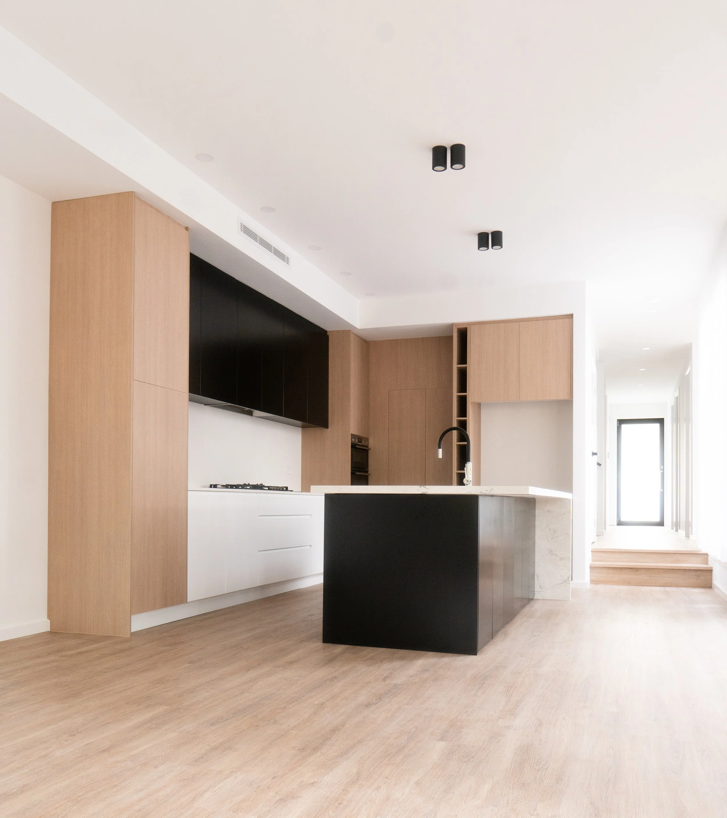 Modern open-concept kitchen with light wood cabinets, black upper cabinets, white lower cabinets, a white marble kitchen island with black sides, and a built-in oven and stove. The space has light wood flooring, black ceiling lights, and a bright hal