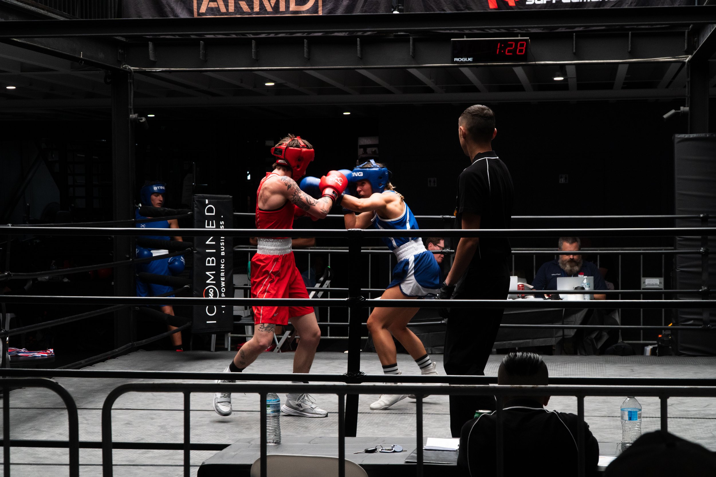 Two female boxers, one in red and one in blue, are fighting inside a boxing ring. A referee is observing the match, and other individuals are seated outside the ring, watching the fight.