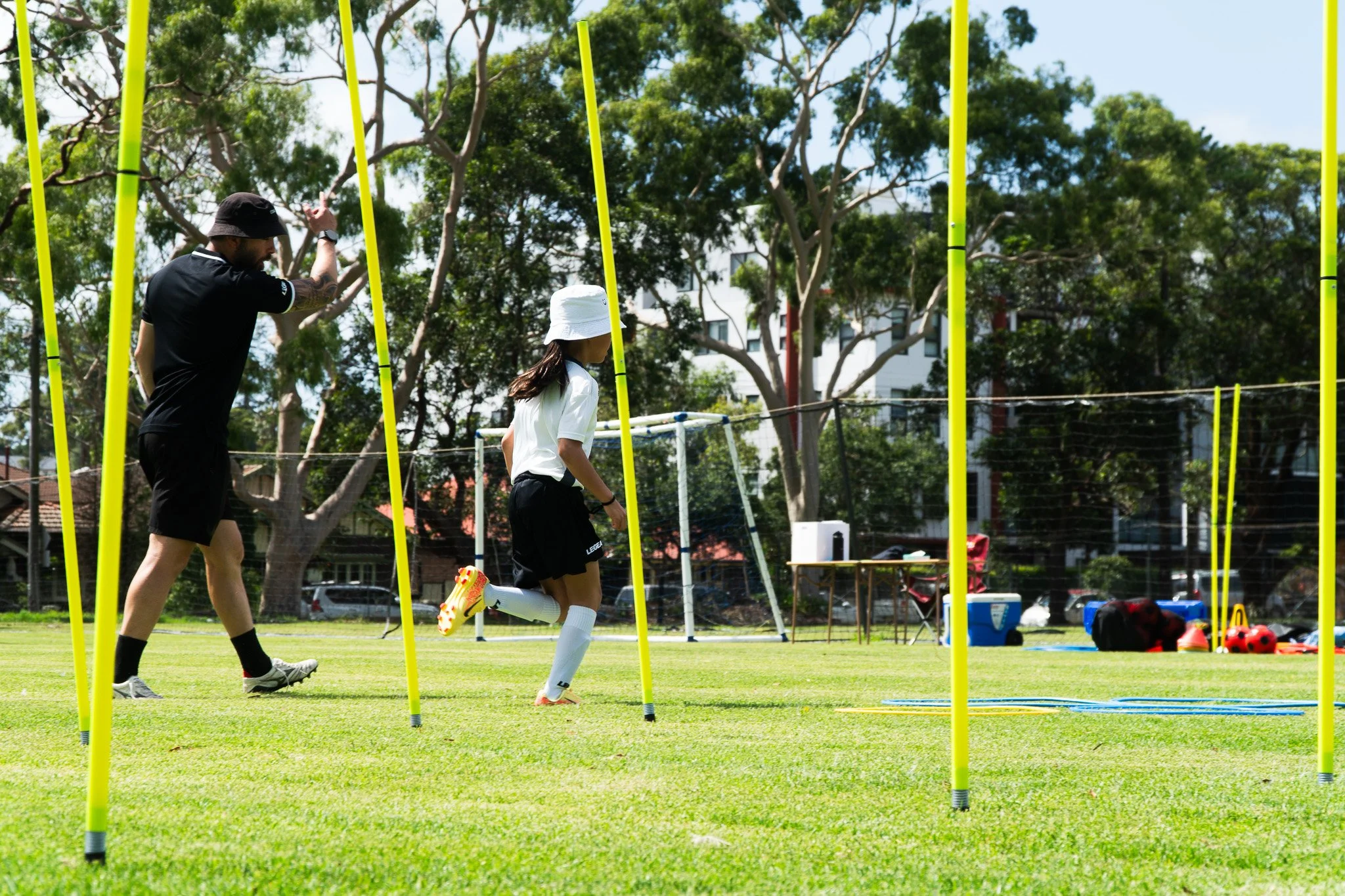 A young girl running through a soccer agility drill course with yellow poles on a grassy field while an instructor watches.