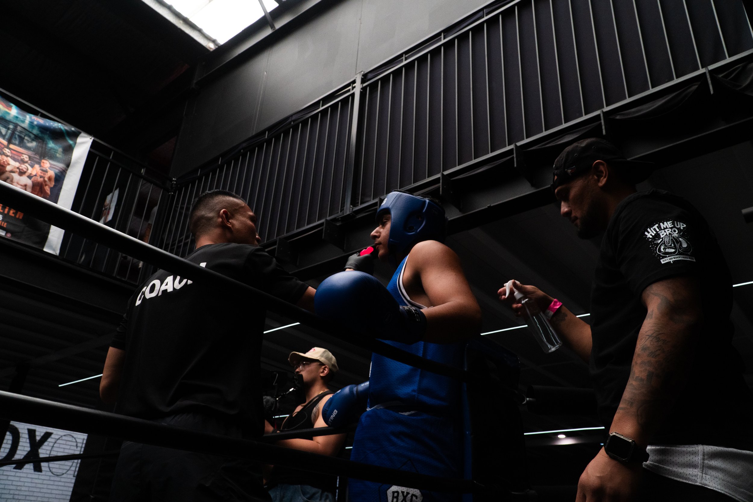 A female boxer wearing blue gear and a helmet in a boxing ring, preparing to compete, with her coach and a support team member nearby.