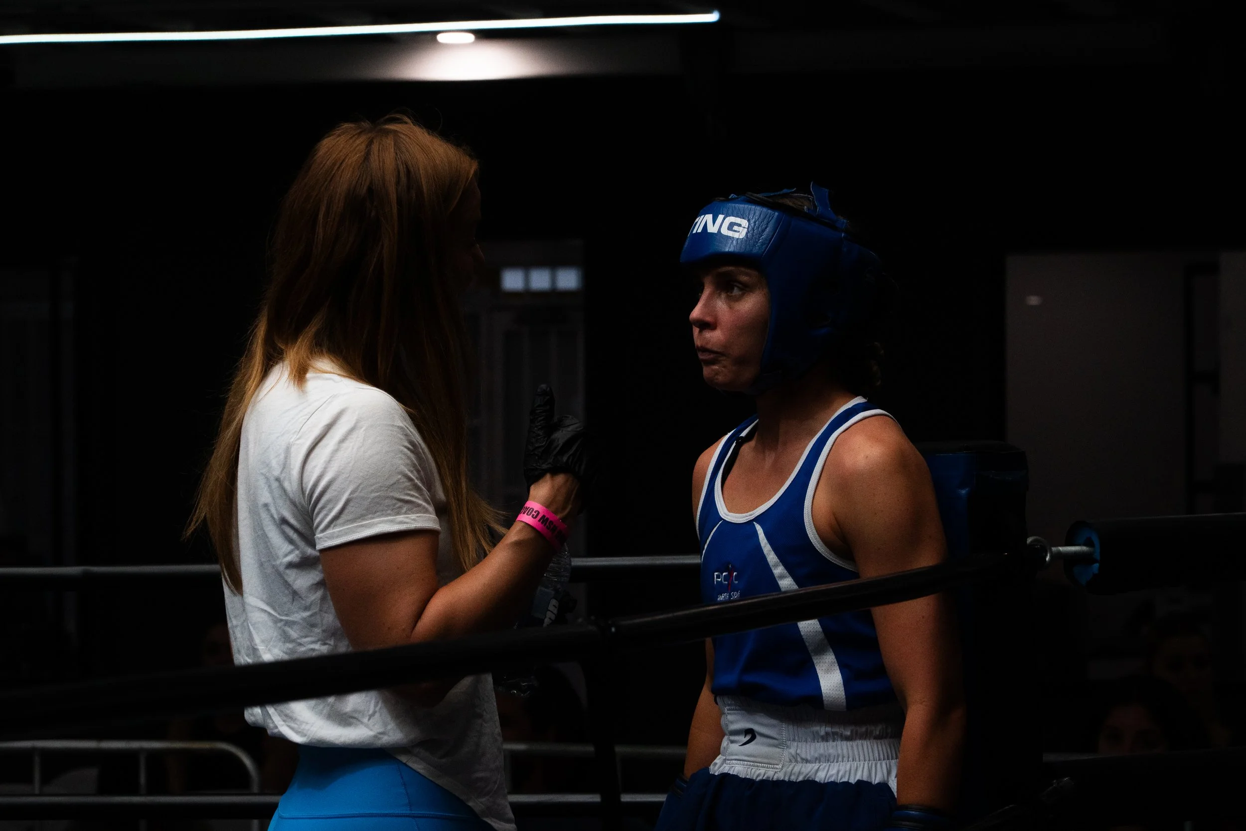 A female boxer wearing a blue helmet and uniform listens to a woman with long brown hair, white t-shirt, and blue pants inside a boxing ring.