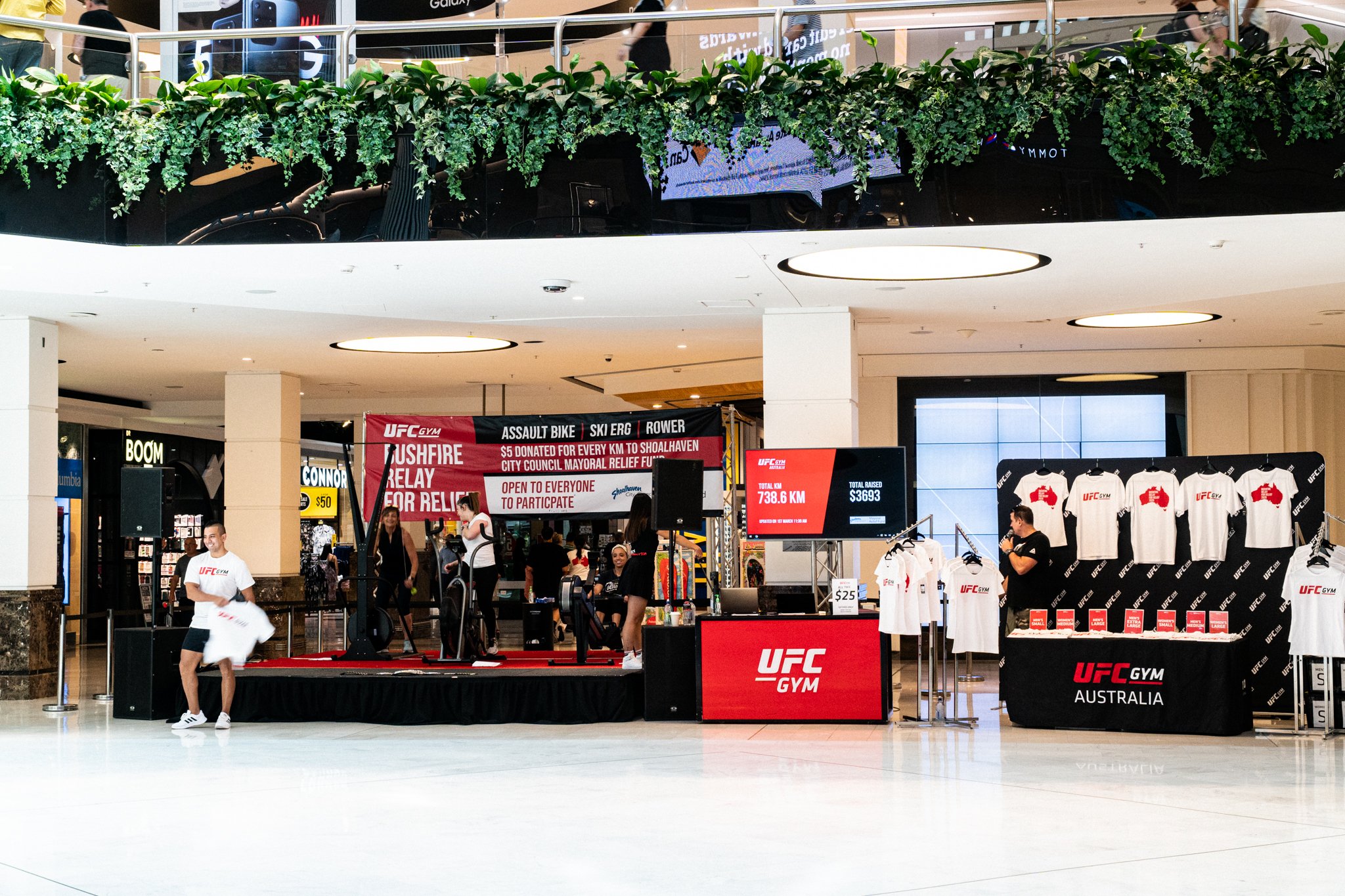 UFC Gym promotional booth inside a shopping mall with a stage and merchandise display. Banners show fundraising details for Shoalhaven City Council relief. A digital screen displays a total of 738.6 km and $3,693 raised. Several people are around, wi