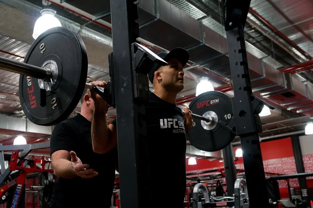 A man doing a squat with a barbell loaded with weight plates at UFC Gym, supervised by a trainer.