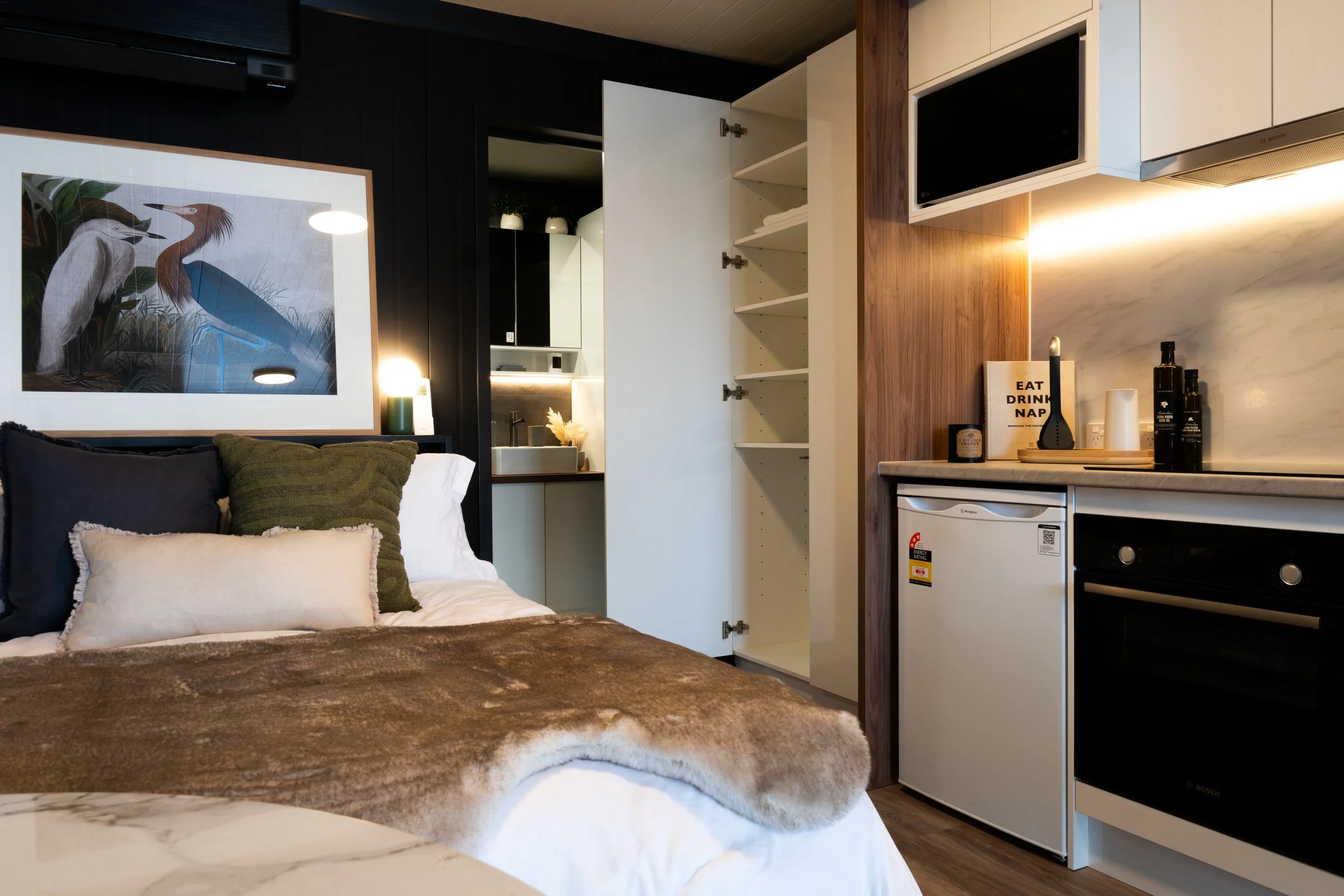 Interior of a modern studio apartment with a bed, a small kitchenette, open closet, and decorative pillows and artwork.