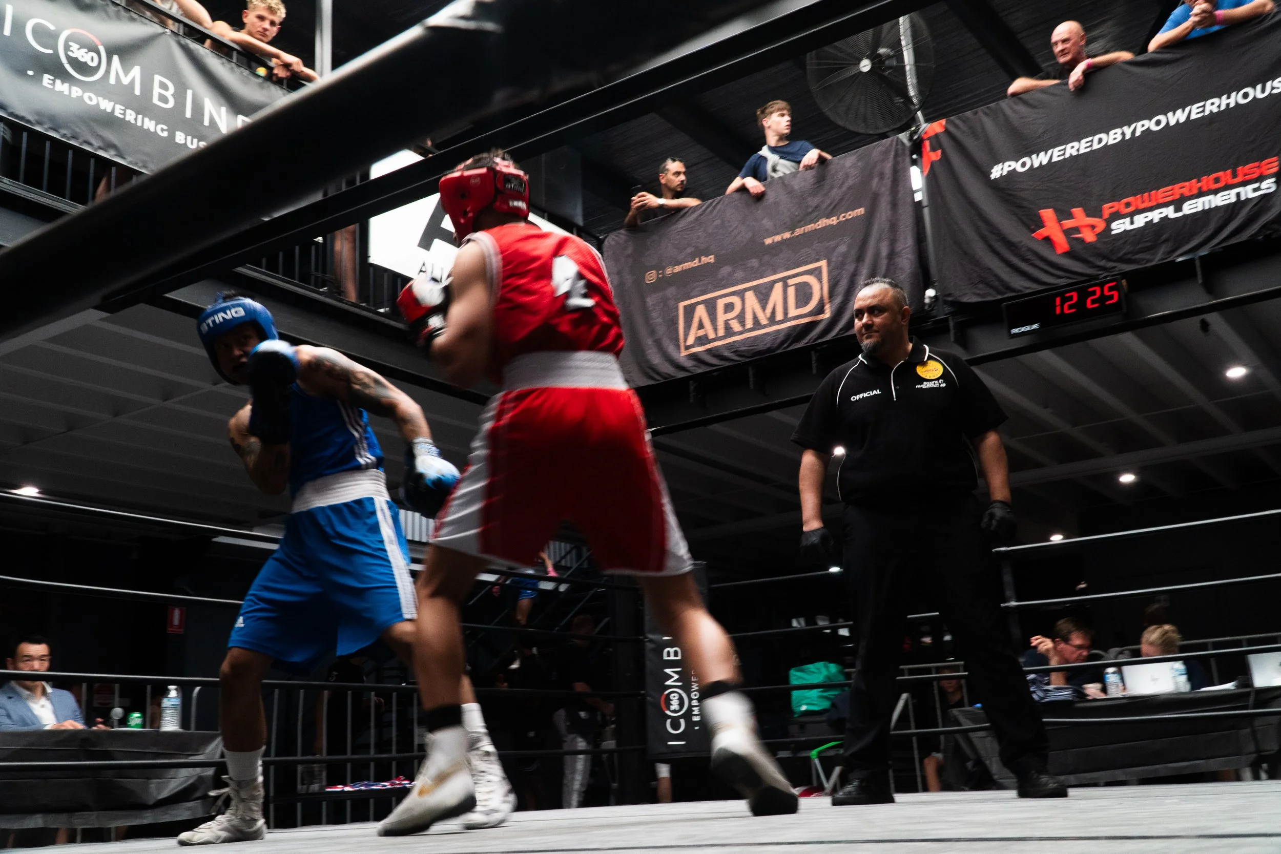 Two boxers in a ring, one in red gear and the other in blue gear, engaged in a match while a referee observes nearby. Spectators watch from the upper level.