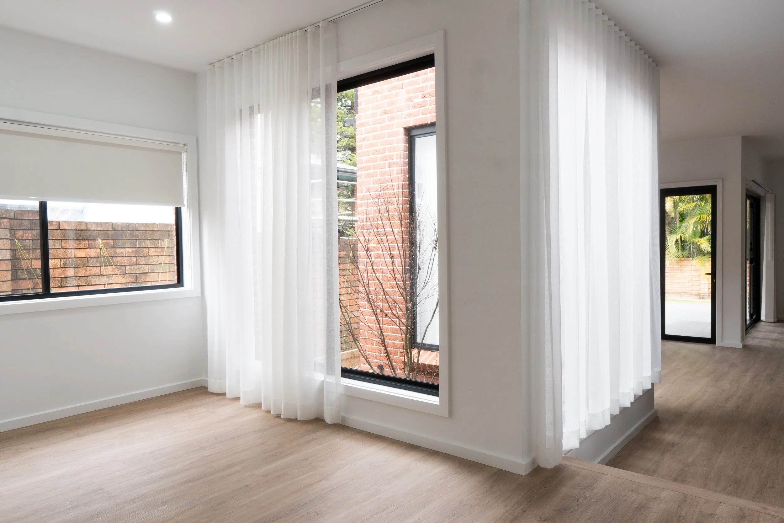 Empty room with white walls, light wood flooring, large windows with white curtains, and a view of a brick wall outside.