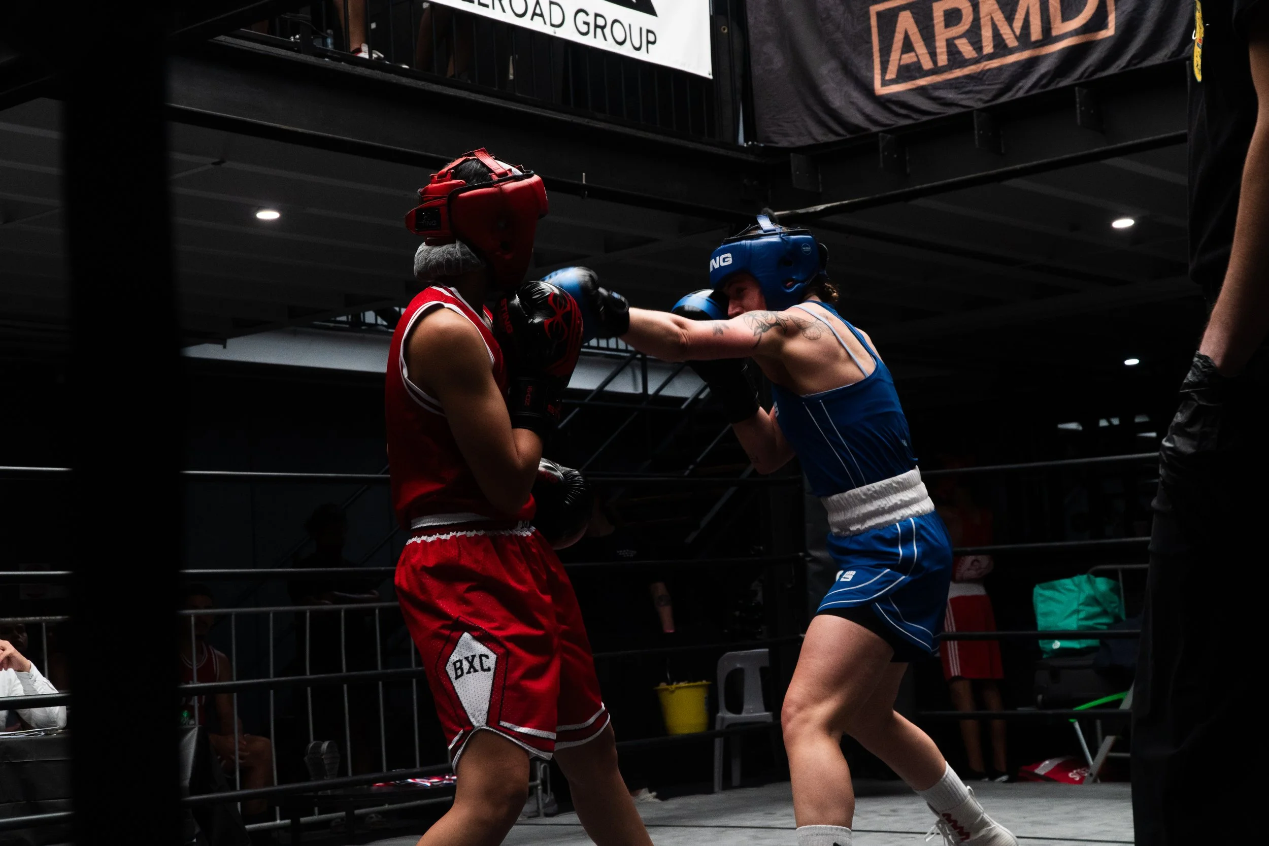 Two female boxers sparring in a boxing ring, one wearing red gear and the other blue gear, with the blue boxer throwing a punch.