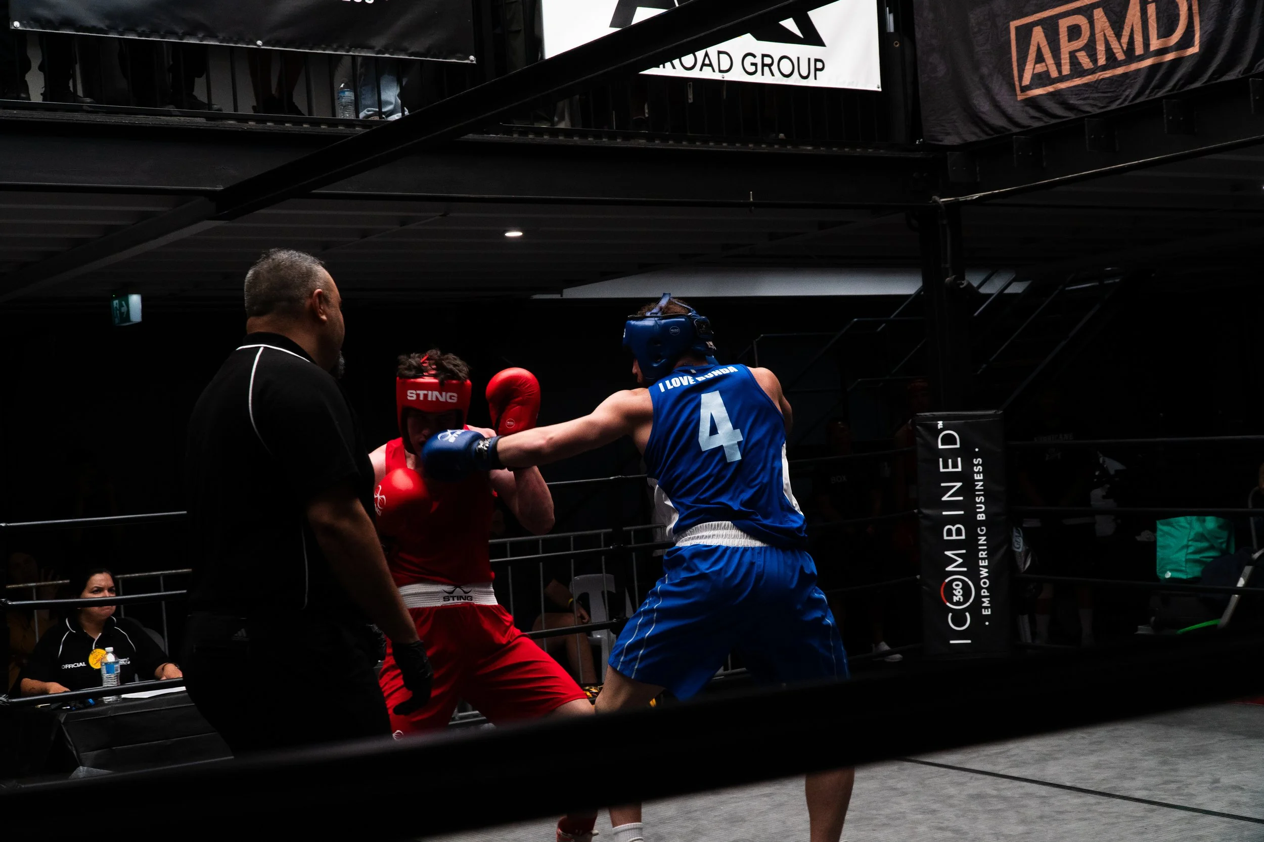 Two boxers, one in red gloves and shorts, the other in blue gloves and shorts, sparring in a boxing ring with a referee watching.