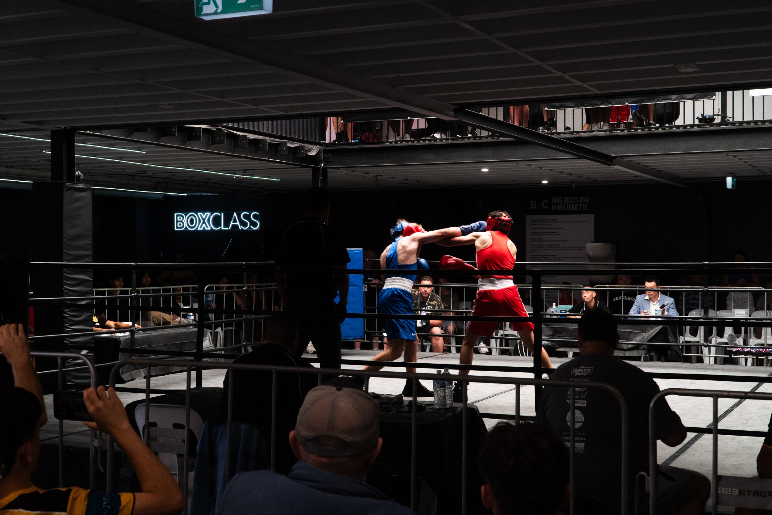 Two boxers in red and blue gear fighting in a boxing ring at a boxing event labeled 'BOXCLASS'. Several spectators are watching, and a referee is inside the ring judging the fight. There is a neon sign in the background that says 'BOXCLASS'.