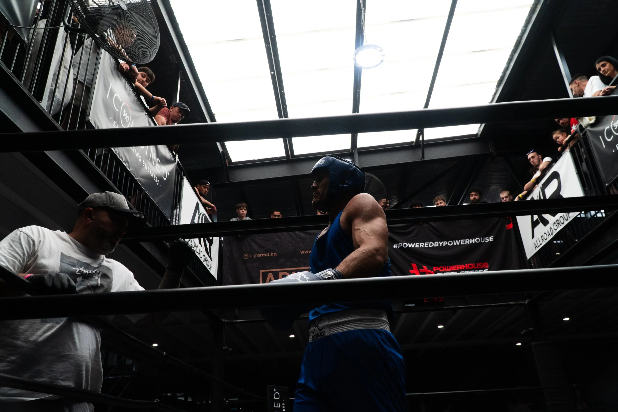 A boxer in blue shorts and a black headgear standing inside a boxing ring, with spectators watching from an upper balcony area.