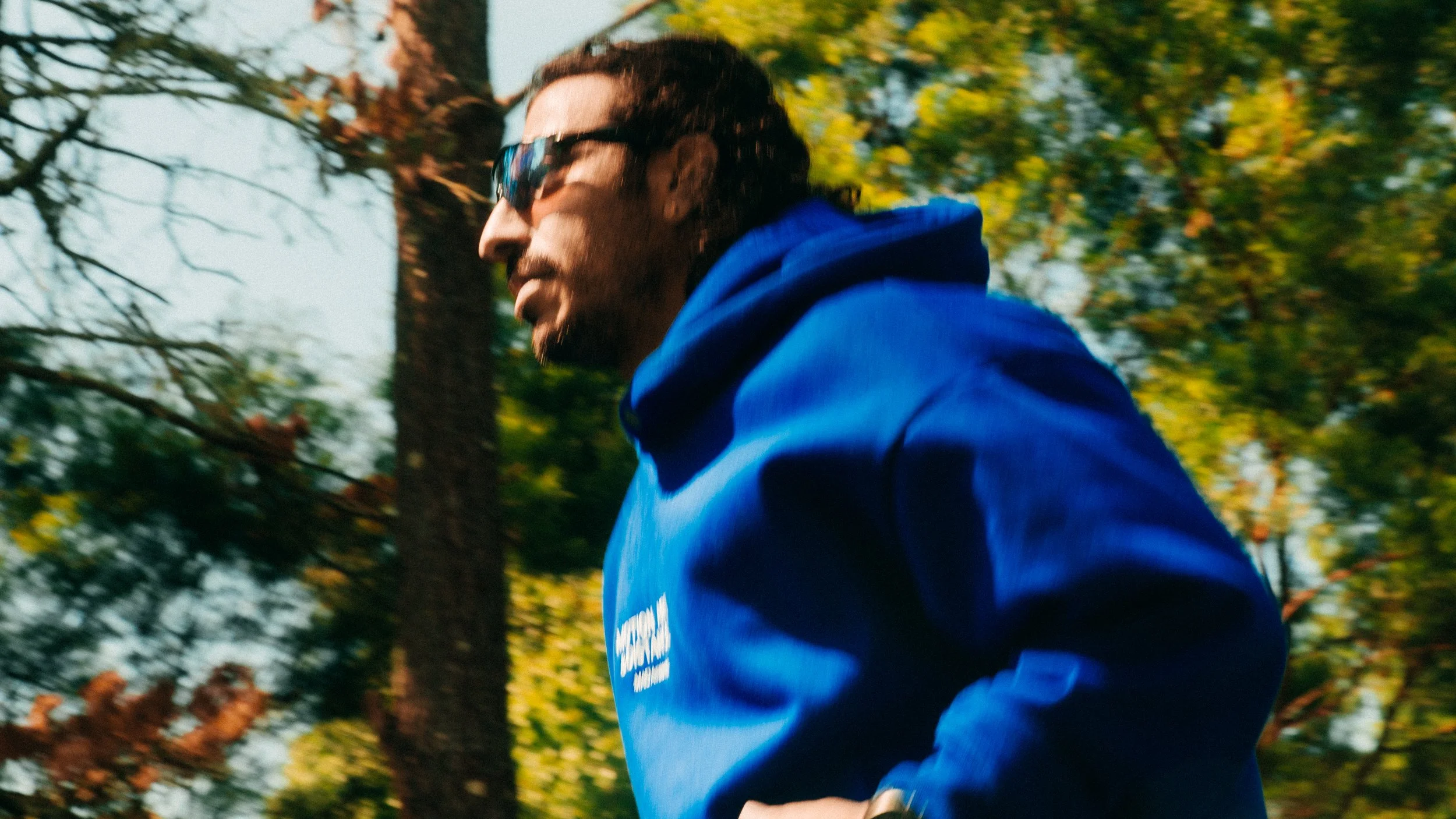 A man wearing sunglasses and a blue hoodie is running outdoors among trees with green and yellow leaves.