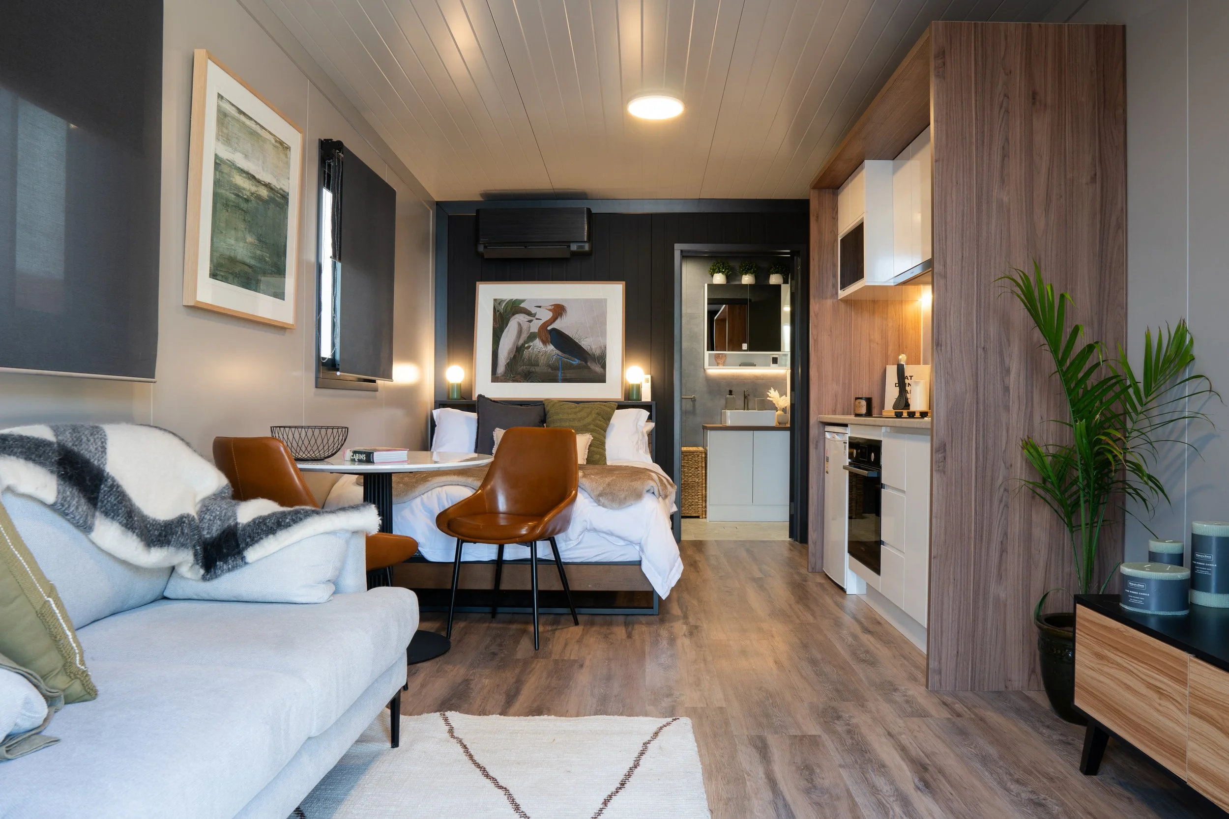 A cozy studio apartment featuring a living area with a white sofa, a small round dining table with two brown chairs, a bed with pillows and a blanket, a kitchenette with white cabinets, a bathroom with a sink, and decorative plants and artwork.