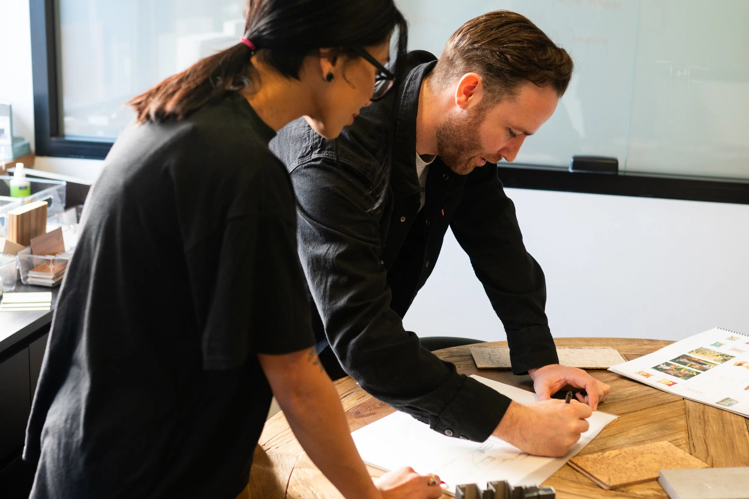 Two people, a woman and a man, are working together on a drawing or design at a round wooden table in an office or studio setting.