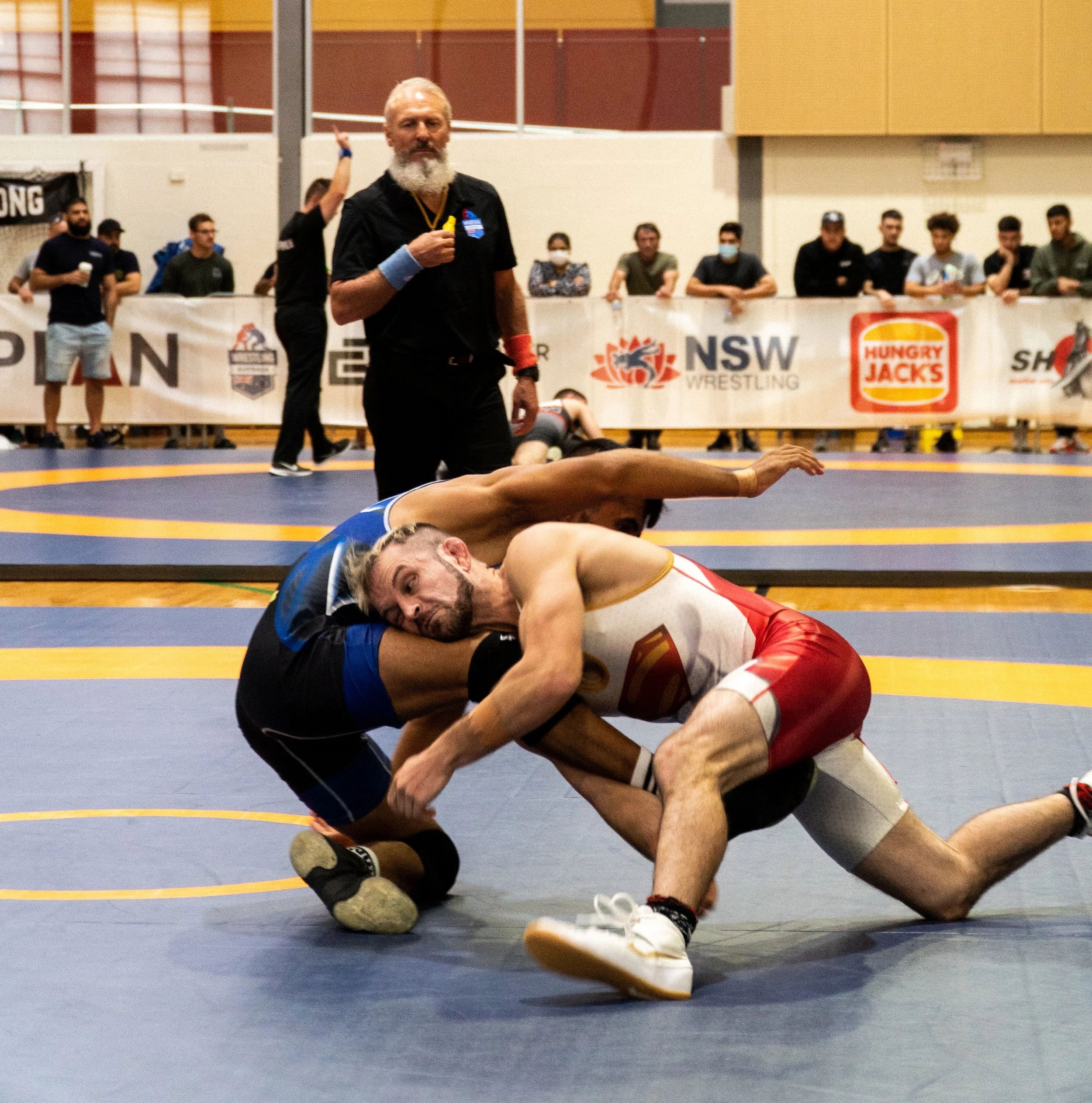 Two male wrestlers on a wrestling mat engaged in a match at a wrestling tournament, with a referee standing behind them and spectators in the background.