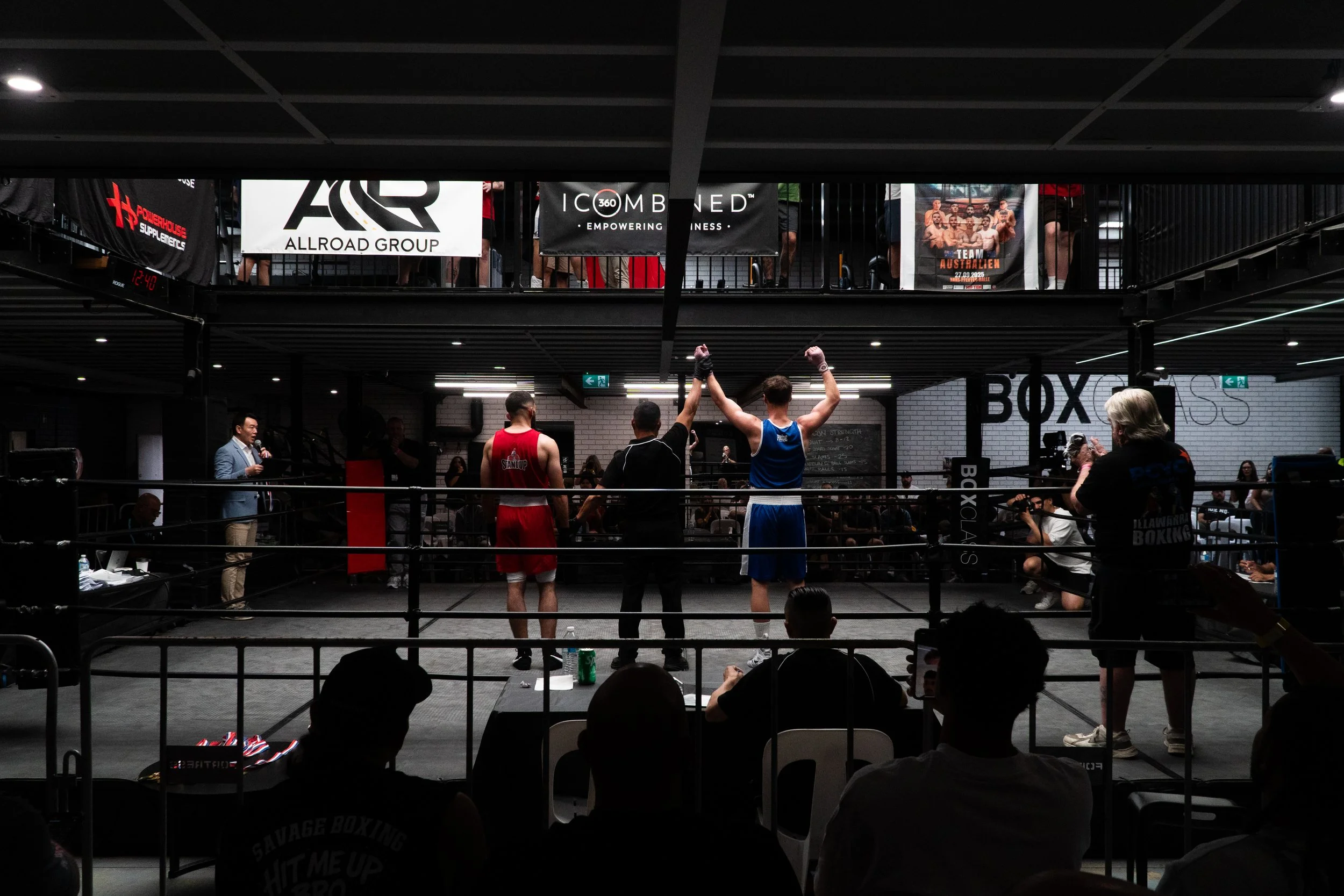 Boxing match with two fighters in the ring, one in red and one in blue, with referee raising the blue fighter's hand in victory, in a gym with spectators and photographers.