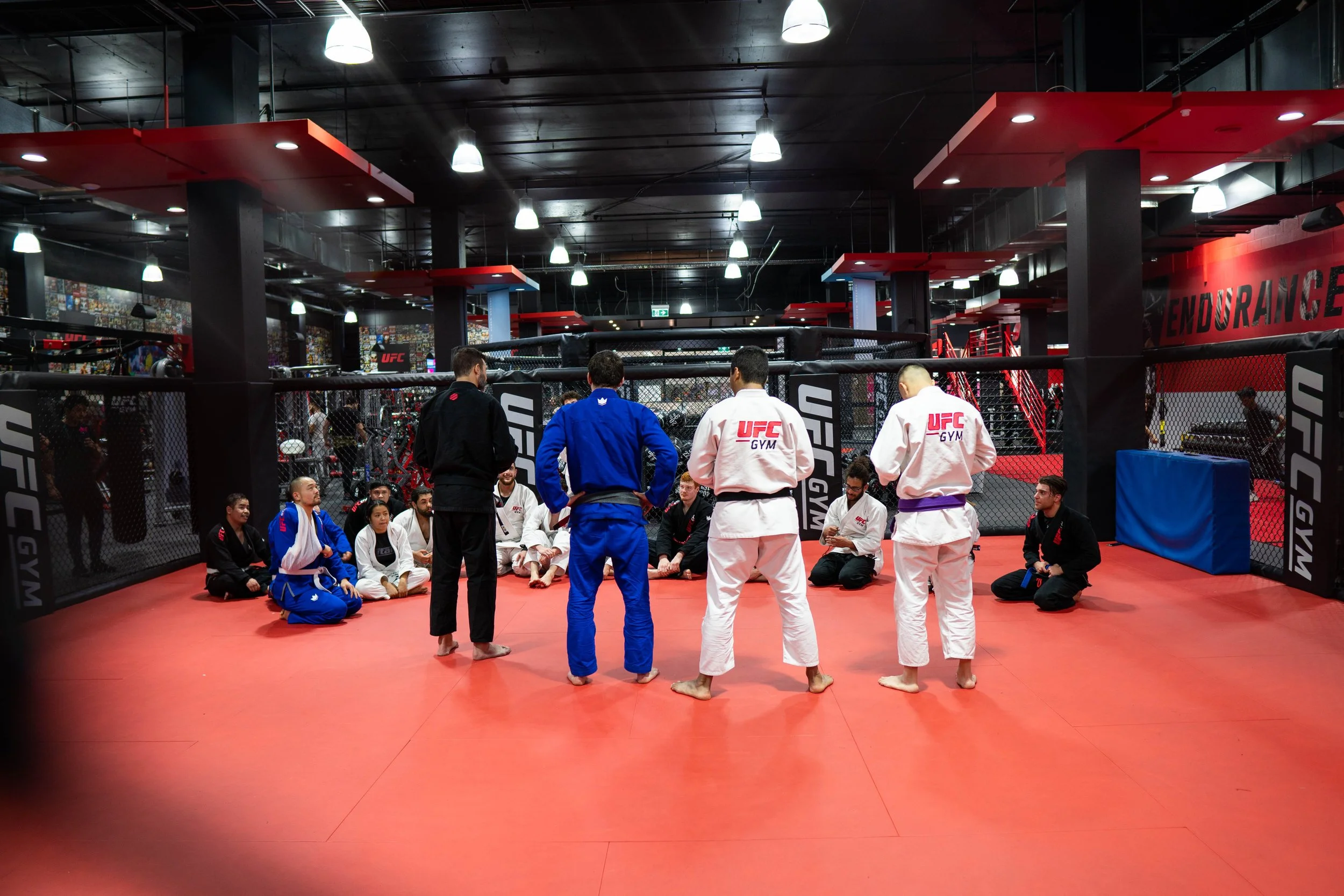 Brazilian Jiu-Jitsu class at UFC Gym with students sitting on the mat and instructor standing in front of them in a training area with UFC branding.