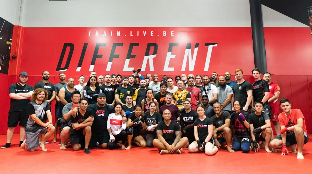 Group of mixed martial artists and athletes gathered inside a gym with a red wall displaying the words "Train. Live. Be different." They are dressed in training gear, some wearing UFC gym apparel, and are posing for a group photo.