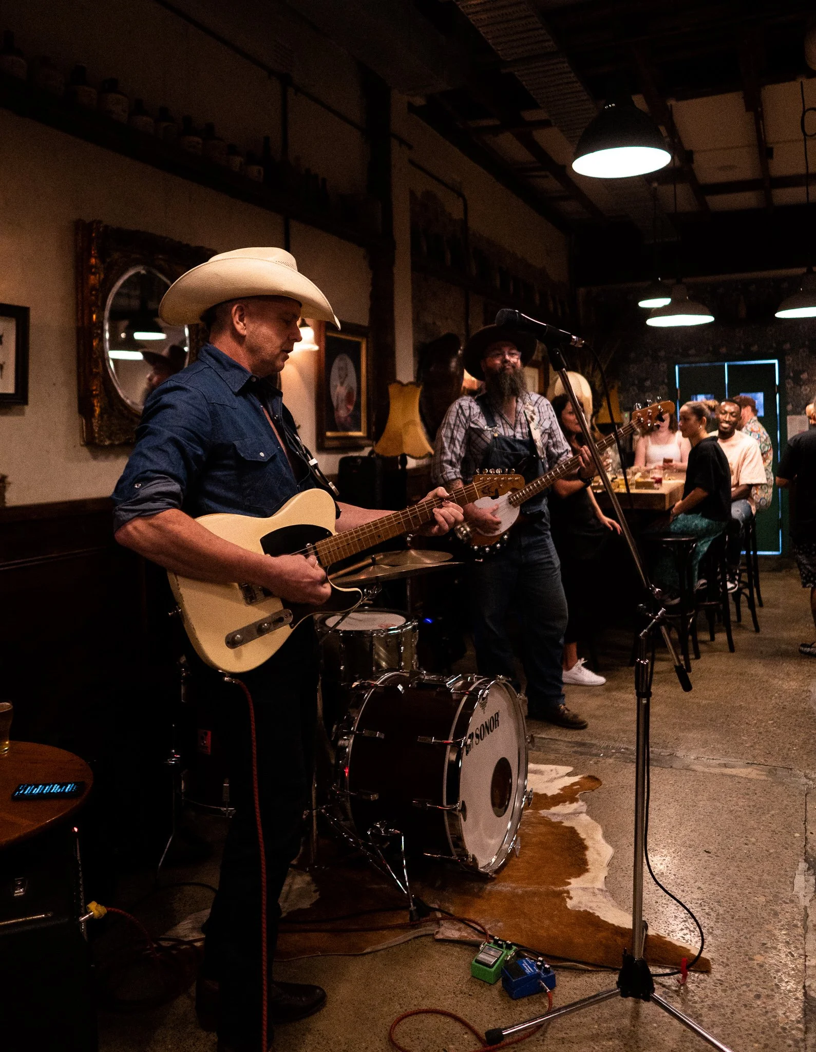 Two musicians performing at a bar, one playing electric guitar and the other playing banjo, with people sitting at the bar and standing around, and a bartender behind the bar.