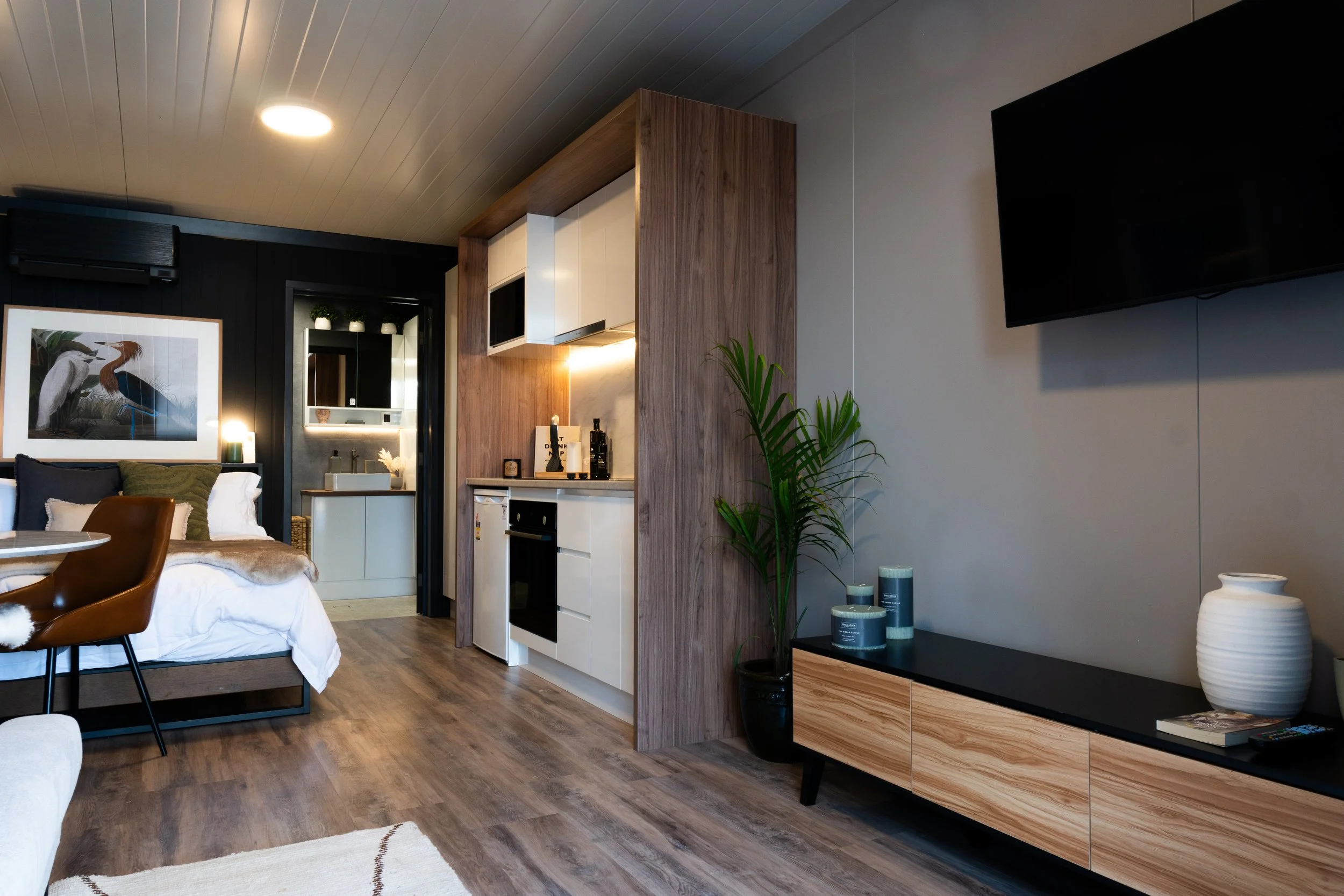 Modern living space with a small kitchenette, a sitting area with a brown chair, and a wall-mounted TV. Decor includes a large framed artwork of a heron, a tall plant, and candles on a black and wood media console.