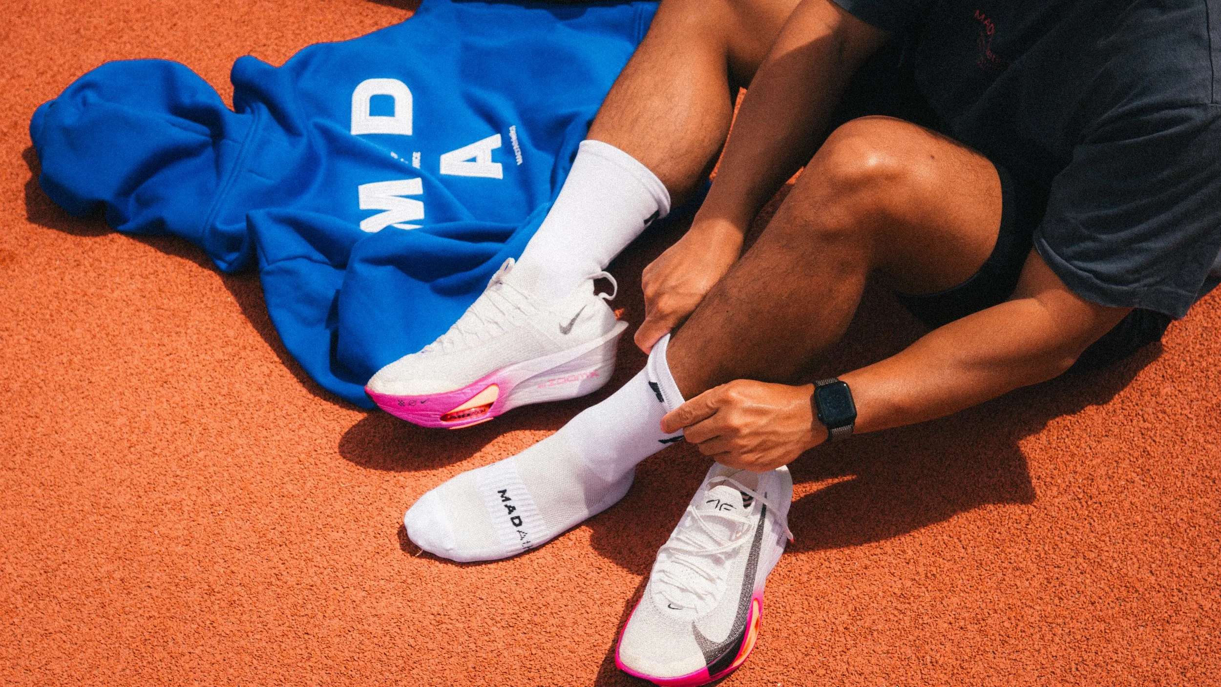 A person is sitting on a track, adjusting their white athletic shoe with pink accents. They are wearing white socks, black shorts, a black shirt, and a black smartwatch. A blue hoodie with white lettering is draped nearby on the red track surface.