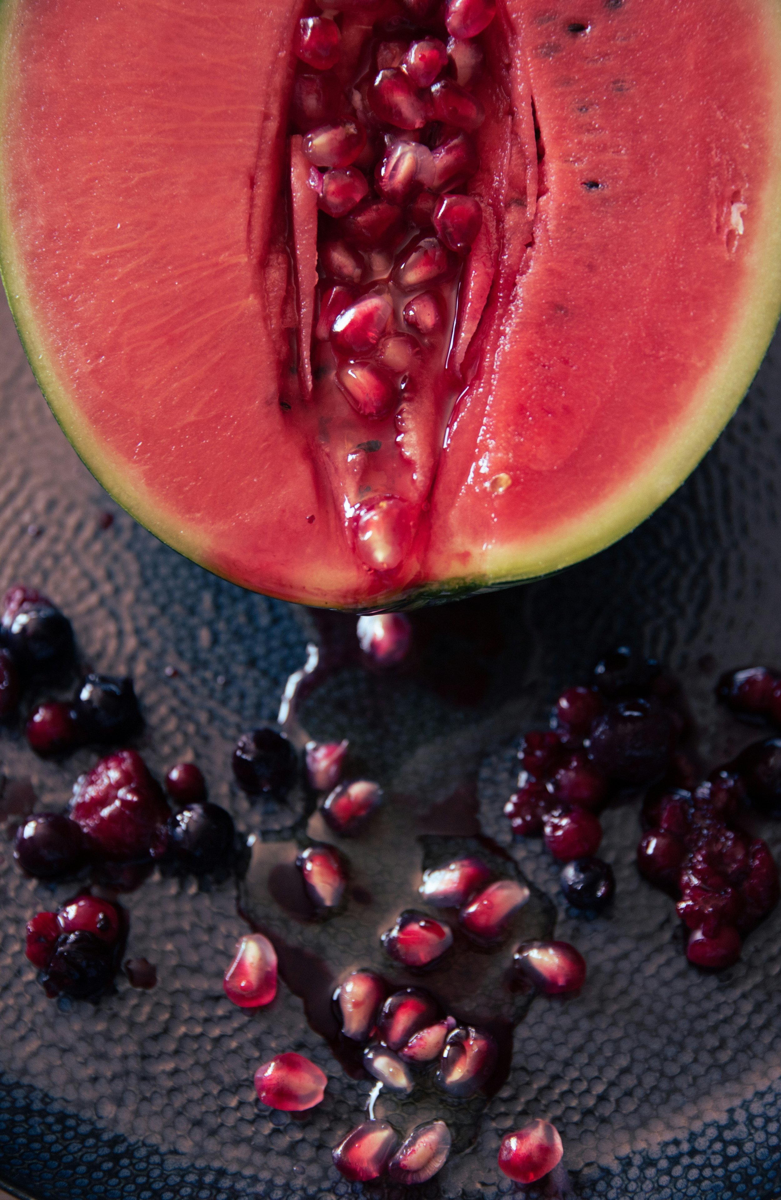 Close-up of a halved pink watermelon with red seeds and black spots, scattered on a black textured surface.