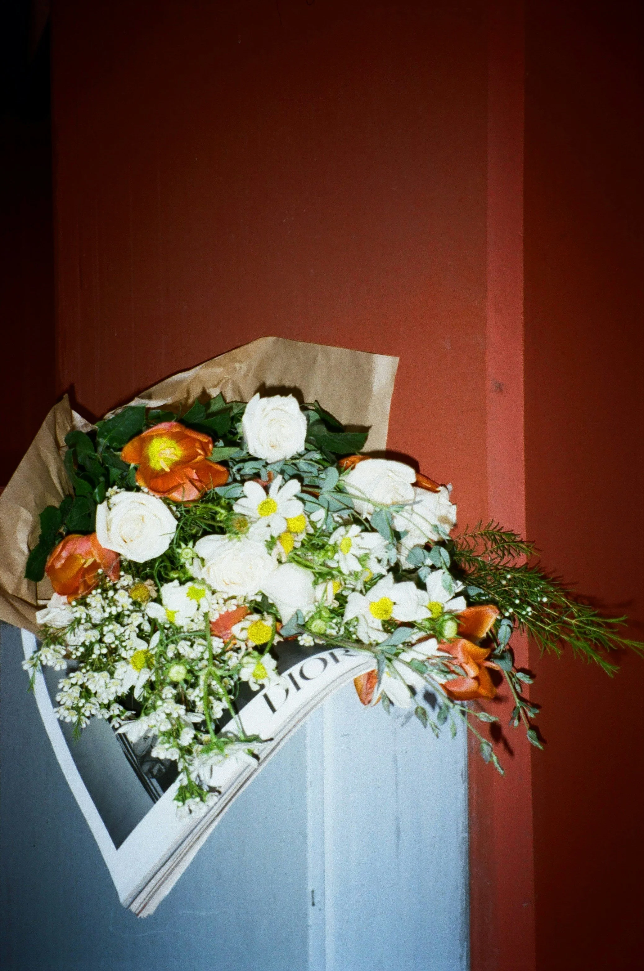 A bouquet of mixed flowers including white roses, orange lilies, and white daisies with greenery in wrapping paper on a table against a red wall.