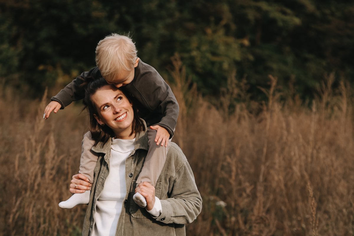 Eine lachende Frau trägt einen kleinen Jungen auf den Schultern in einem Feld bei Sonnenuntergang.