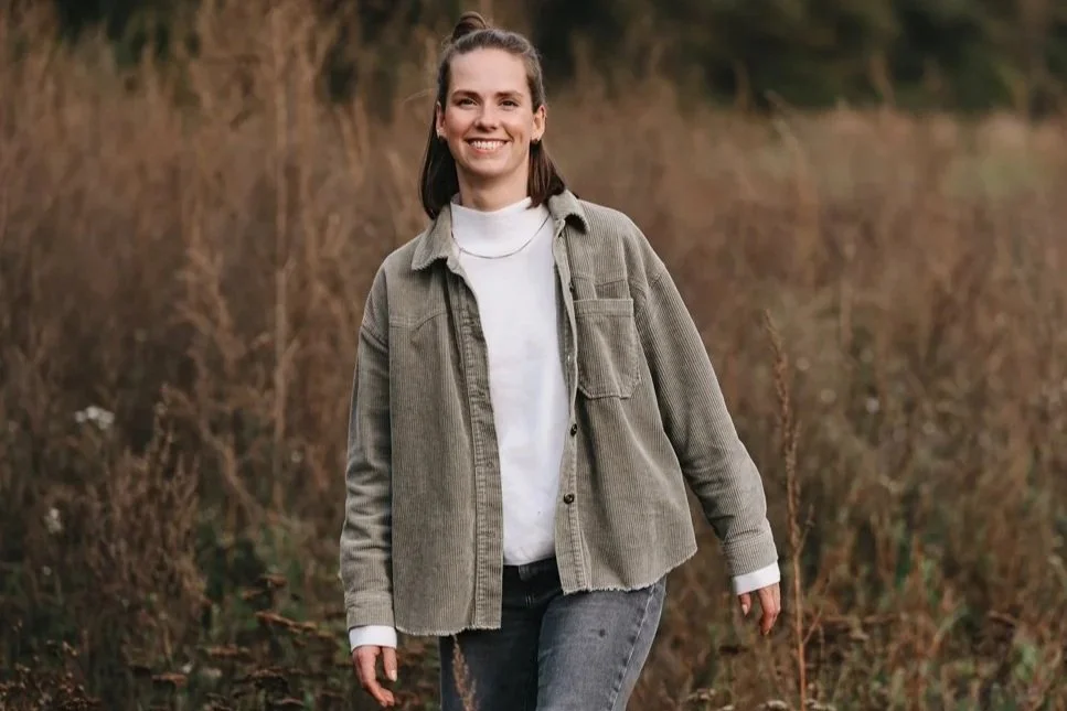 Eine junge Frau in einem Feld, lächelnd, trägt eine beige Jacke und eine weiße Bluse.