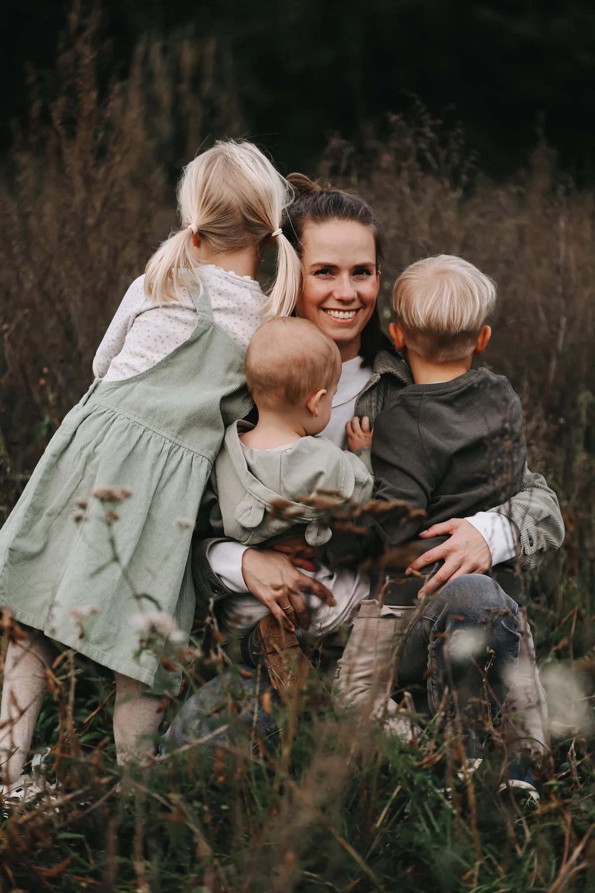 Eine Frau umarmt drei kleine Kinder im Freien, umgeben von Pflanzen und Bäumen, lächelt glücklich.