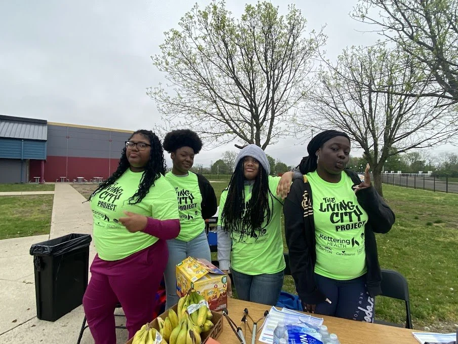 Four young women standing outdoors behind a table with bananas, juice boxes, and bottled water, wearing bright green shirts with 'The Living City Project' printed on them, at a community event or project in a park.