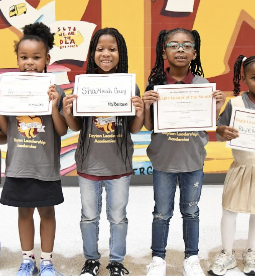 Four young girls standing side-by-side, holding awards and certificates, in a colorful school setting. They are wearing gray shirts with a logo that says 'Dream Leadership Academy,' and are smiling.