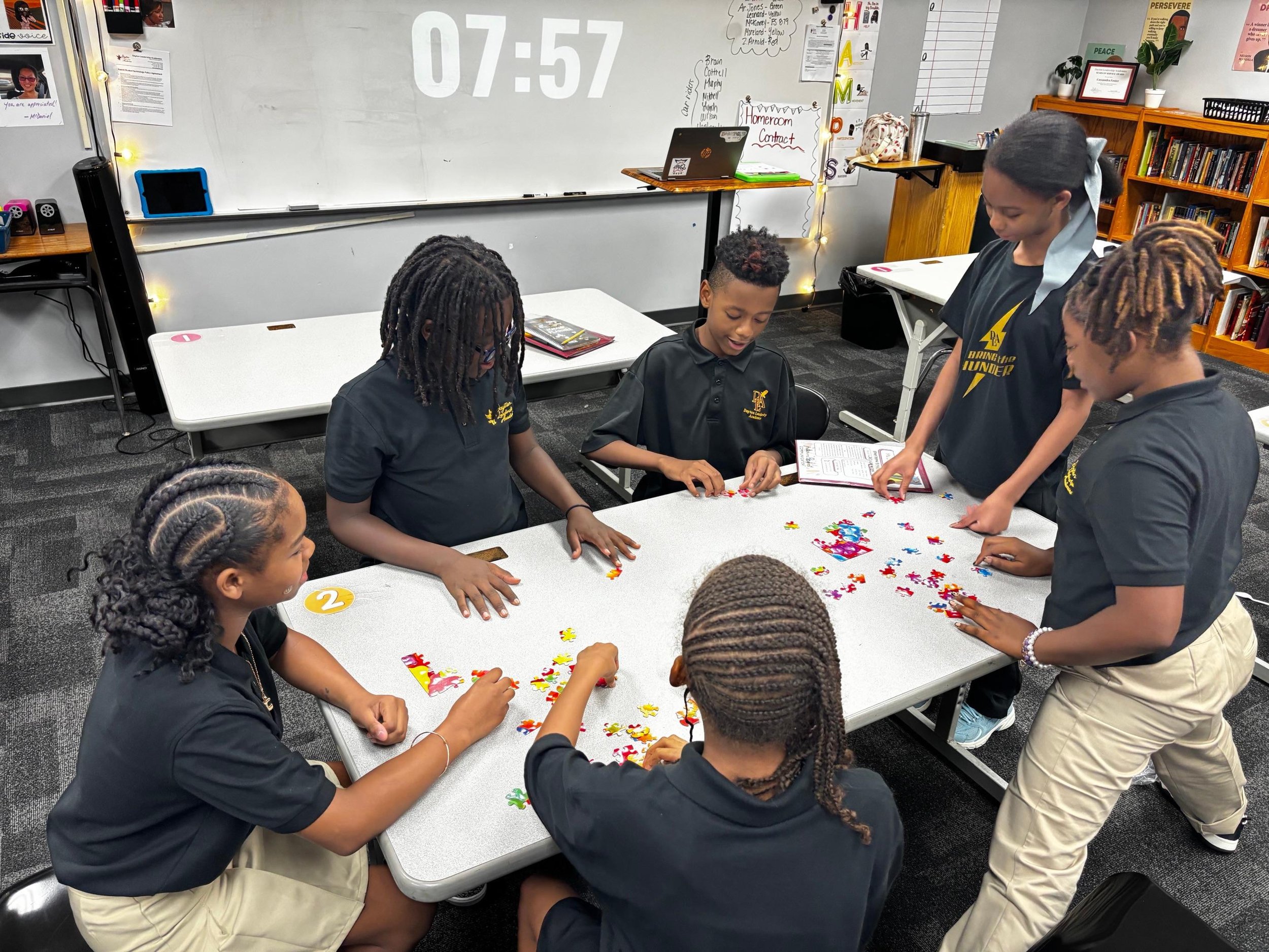 Six students working on jigsaw puzzle at a classroom table, with a whiteboard behind them showing the time 07:57, and shelves with books and classroom decorations in the background.