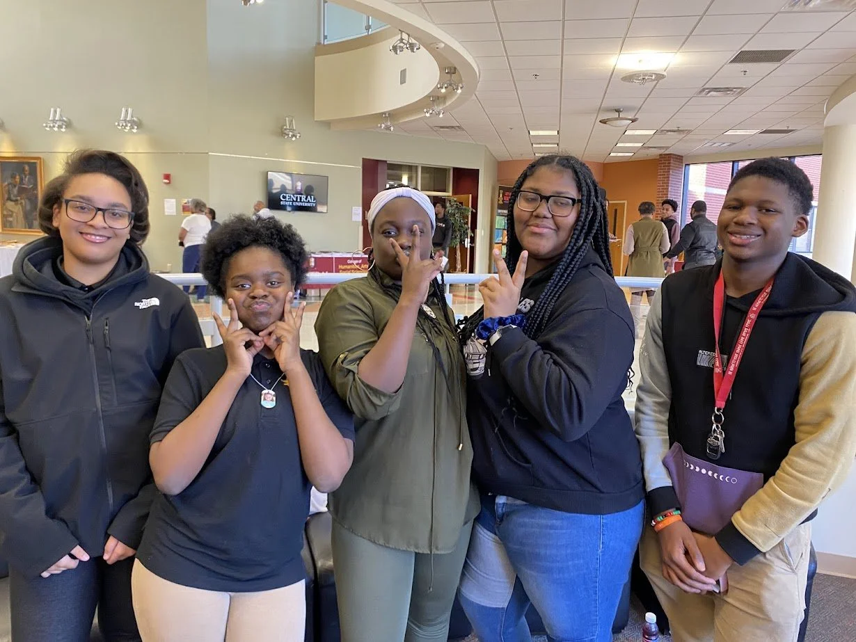 Group of five young people standing together indoors, smiling, making peace signs, wearing casual clothing and jackets.