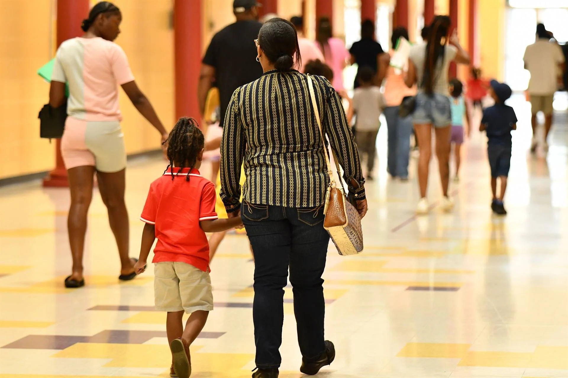A woman and a young girl holding hands as they walk through a crowded indoor space with colorful walls and multiple people, including children and adults, in the background.