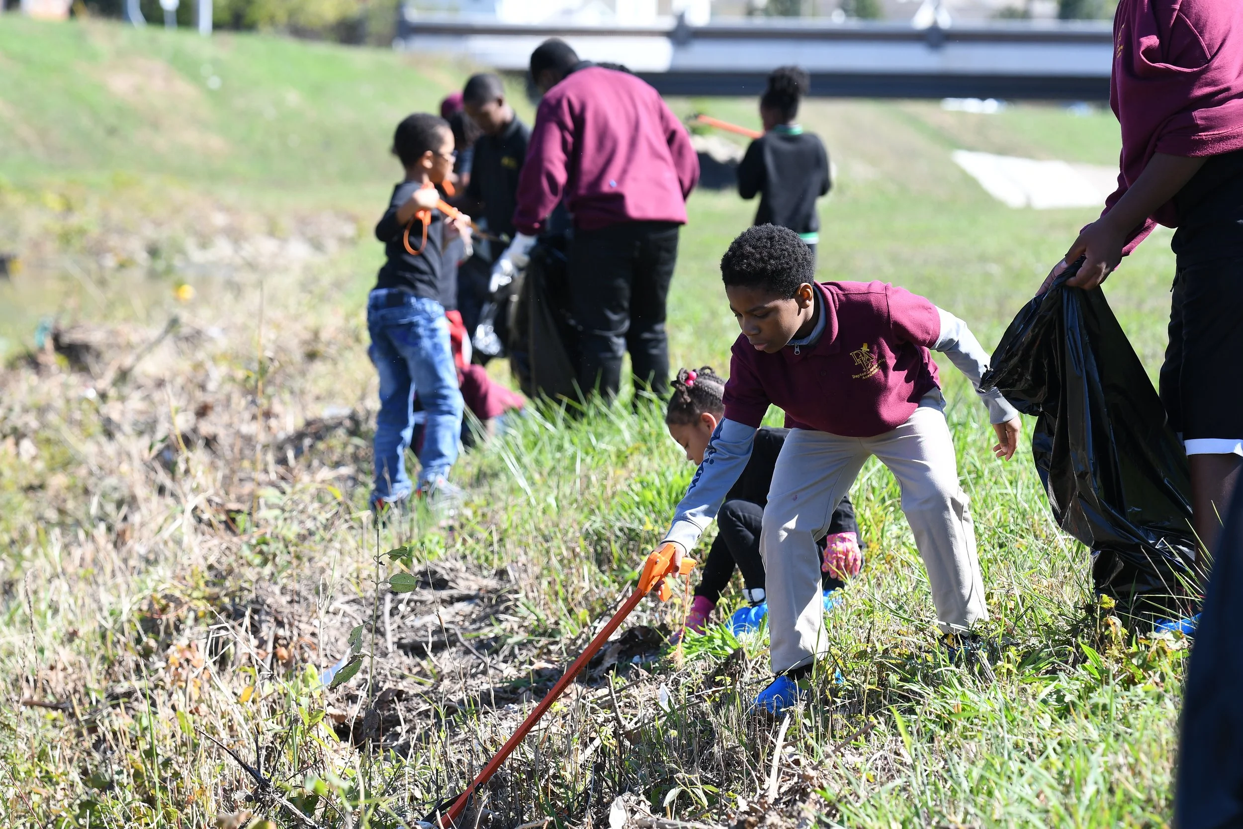 Children and adults participating in a community cleanup, collecting trash from a grassy hillside.