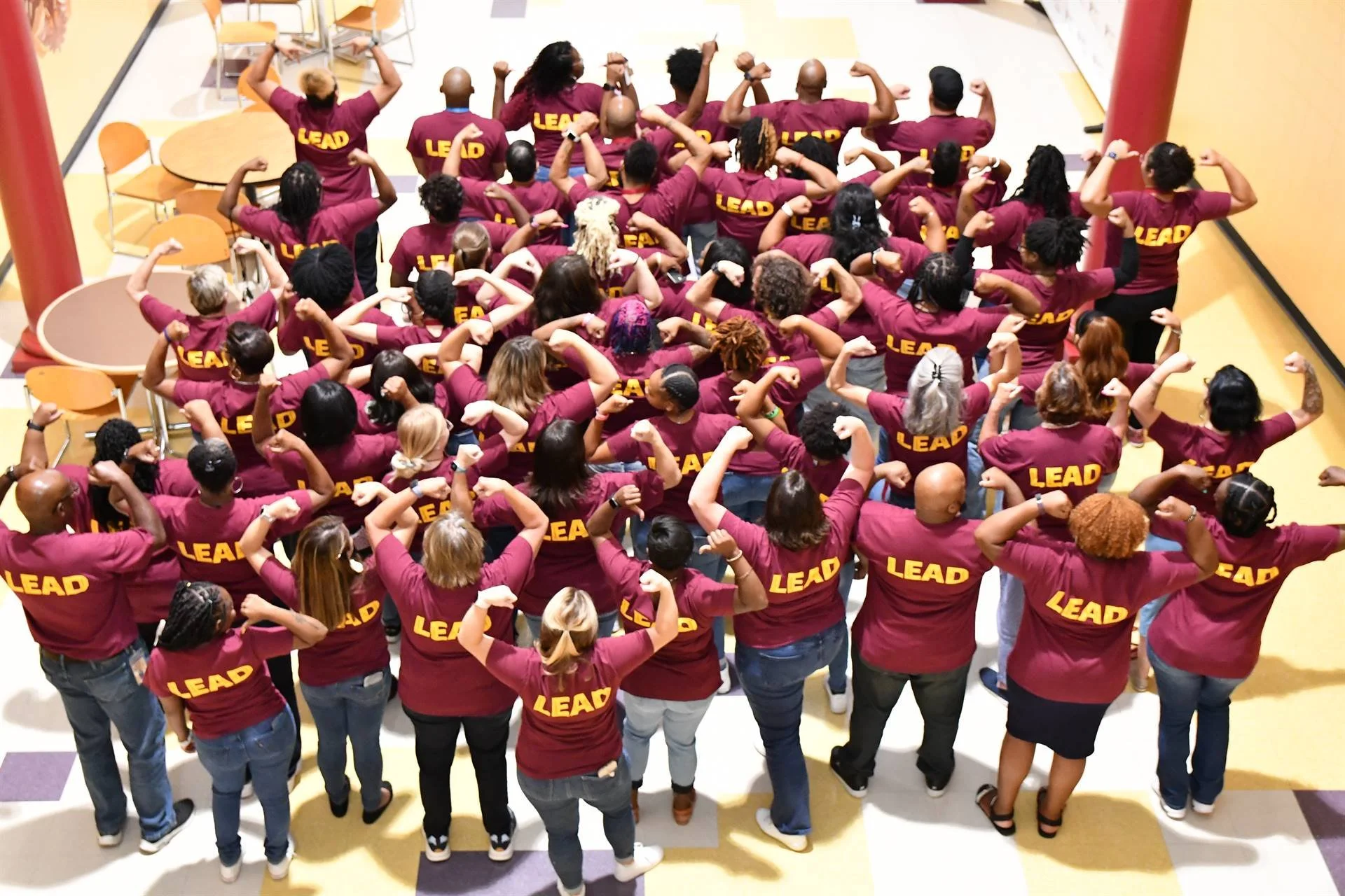 A group of people wearing matching maroon shirts with 'LEAD' printed on the back, standing in a large room, flexing their arm muscles in a display of strength and unity.