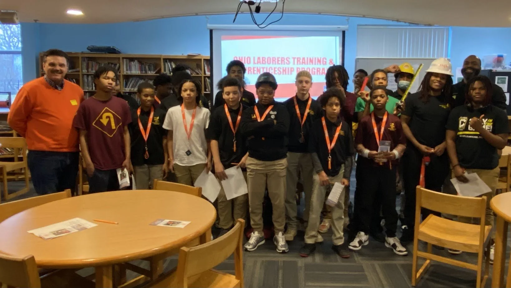 A group of students and adults gathered in a library or classroom for a training and apprenticeship program. There is a screen in the background that says 'Ohio Laborers Training & Apprenticeship Program.' The group includes diverse individuals, some wearing hard hats and safety vests, and they are posing for a group photo.