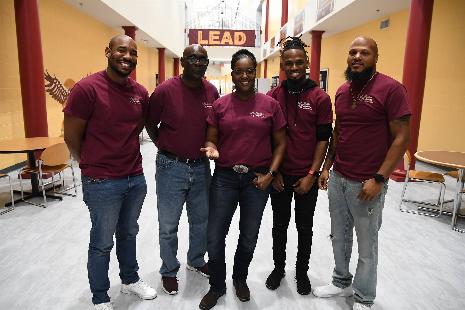 Group of five people standing together indoors, all wearing burgundy shirts, smiling at the camera.