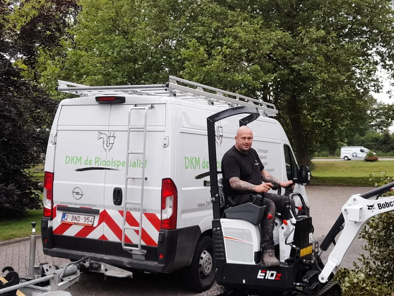 Monteur van DKM de Rioolspecialist op een minigraafmachine naast de witte bedrijfsbus met groene belettering, klaar voor rioolwerk in Breda en omgeving.