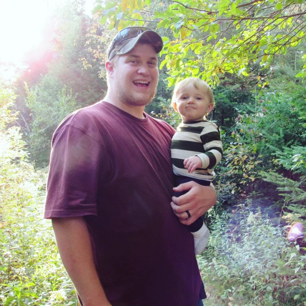 A man holding a young child outdoors in a lush green forest with sunlight filtering through the trees.