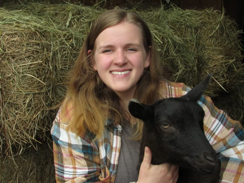 A young woman with long, light brown hair smiling while holding a black goat in front of a hay bales.