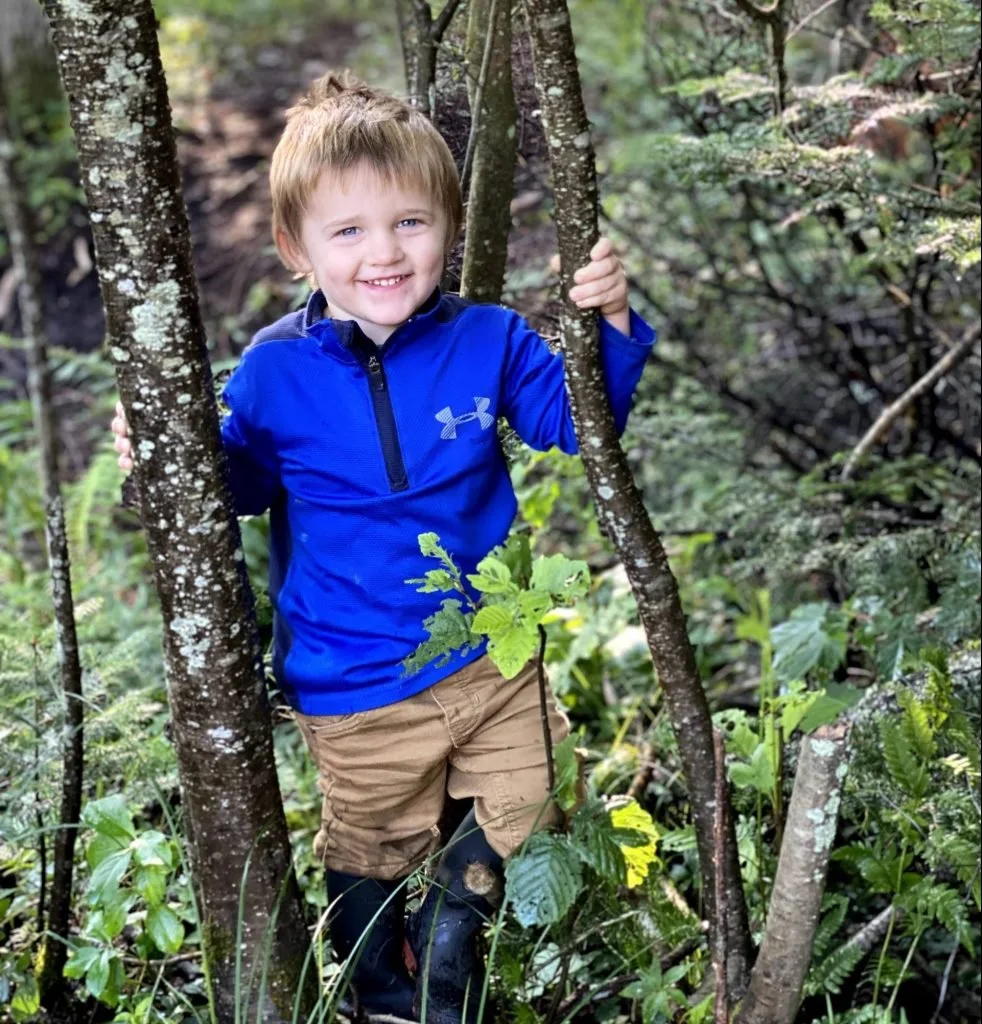 A young boy with blonde hair and blue eyes smiling while standing among trees in a forest, holding onto a tree with one hand, wearing a blue jacket, tan shorts, and black rubber boots.