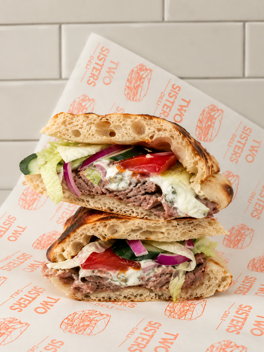 A sandwich with arugula and red peppers, placed on branded paper with a white tiled wall in the background.