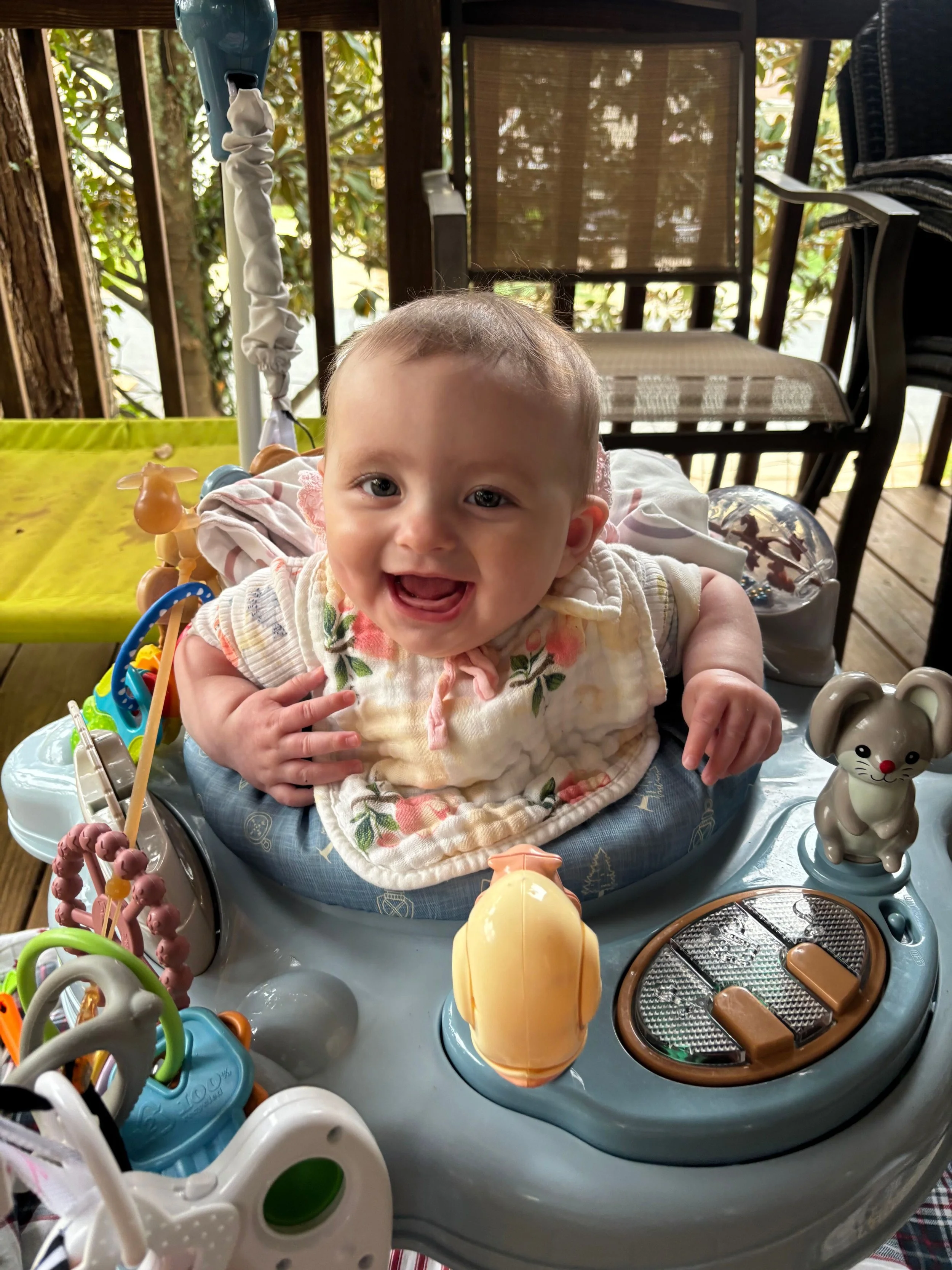 A smiling baby sitting in a baby walker on a porch, surrounded by toys and a toy mouse figurine.